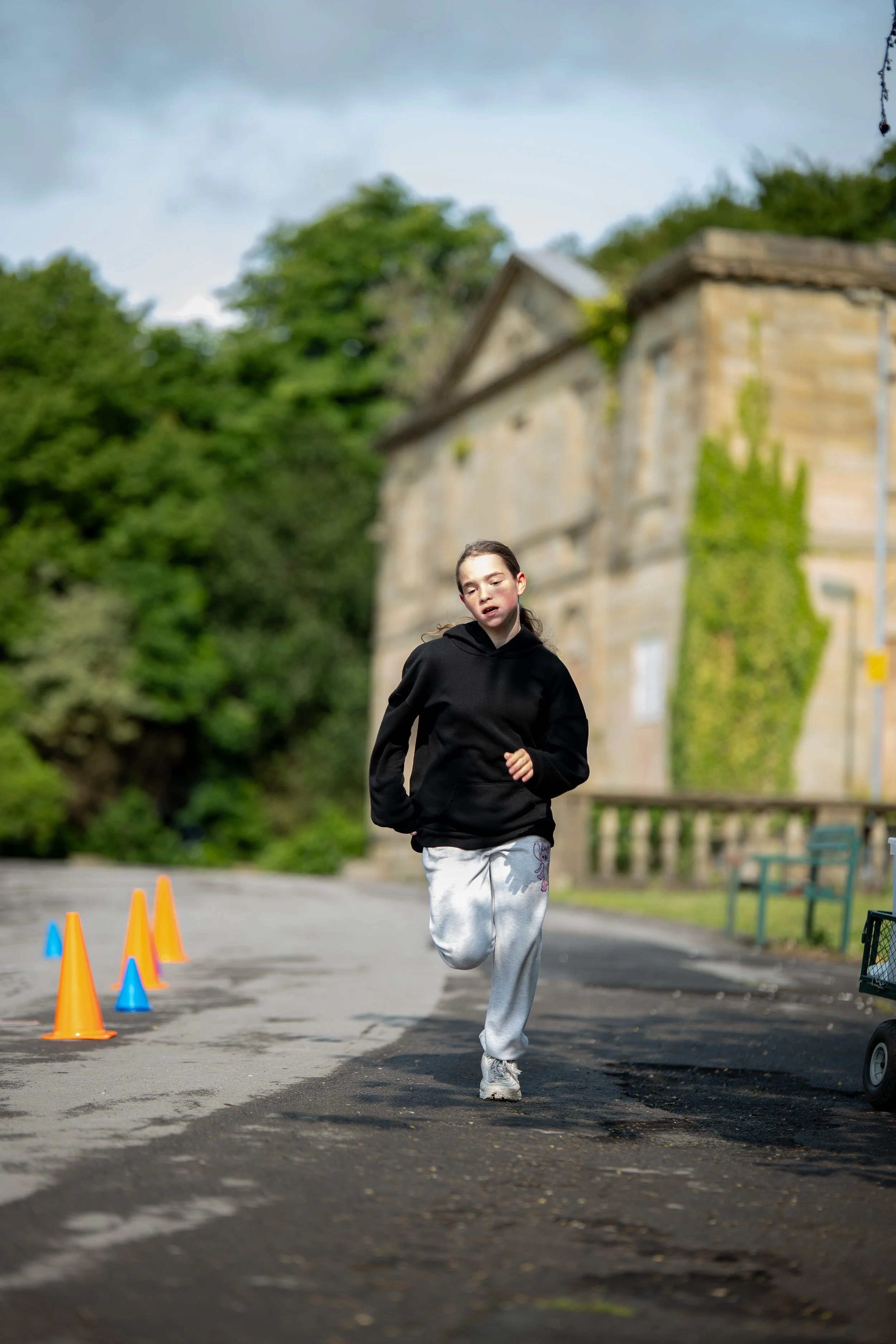 A young girl running outdoors along a paved path, wearing a black hoodie and gray sweatpants, with orange and blue cones set up on the path, and an old stone building with greenery behind her.