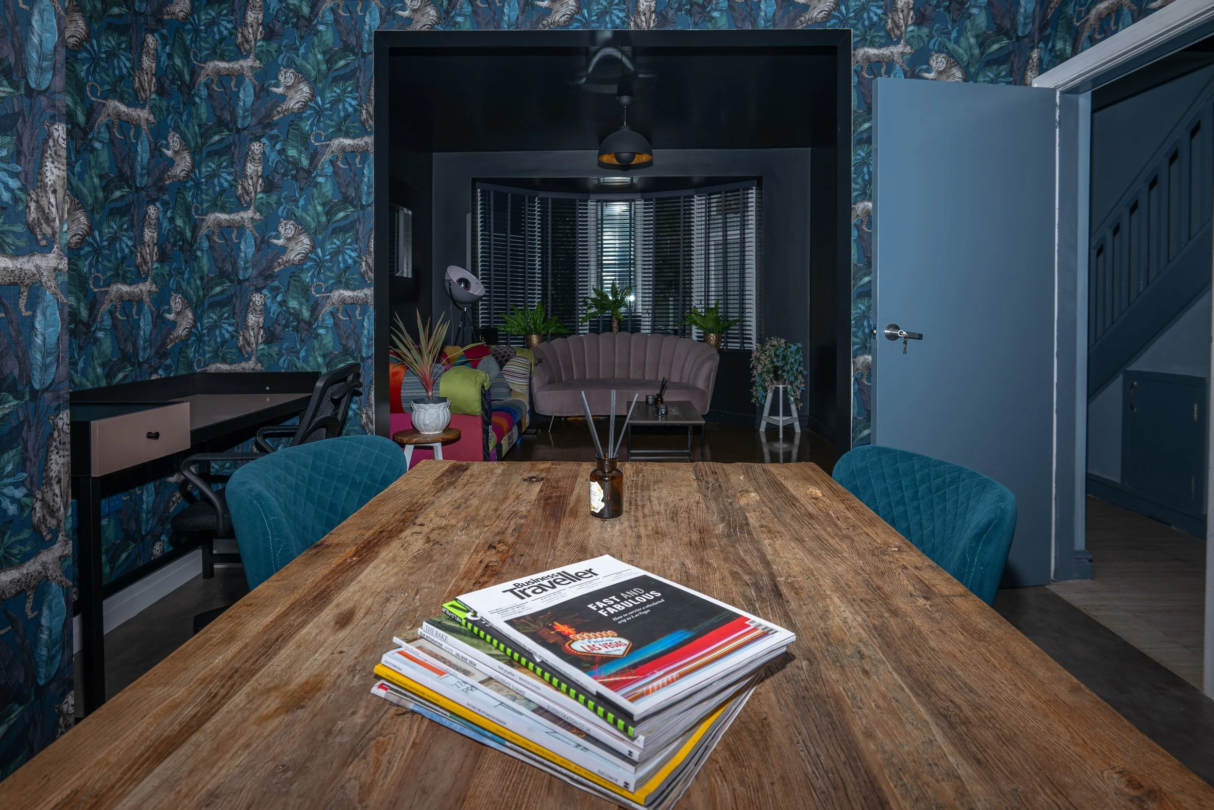 A wooden dining table with a stack of magazines, surrounded by teal upholstered chairs, in a room with dark walls, tropical wallpaper, and a separate sitting area with colorful furniture and plants.