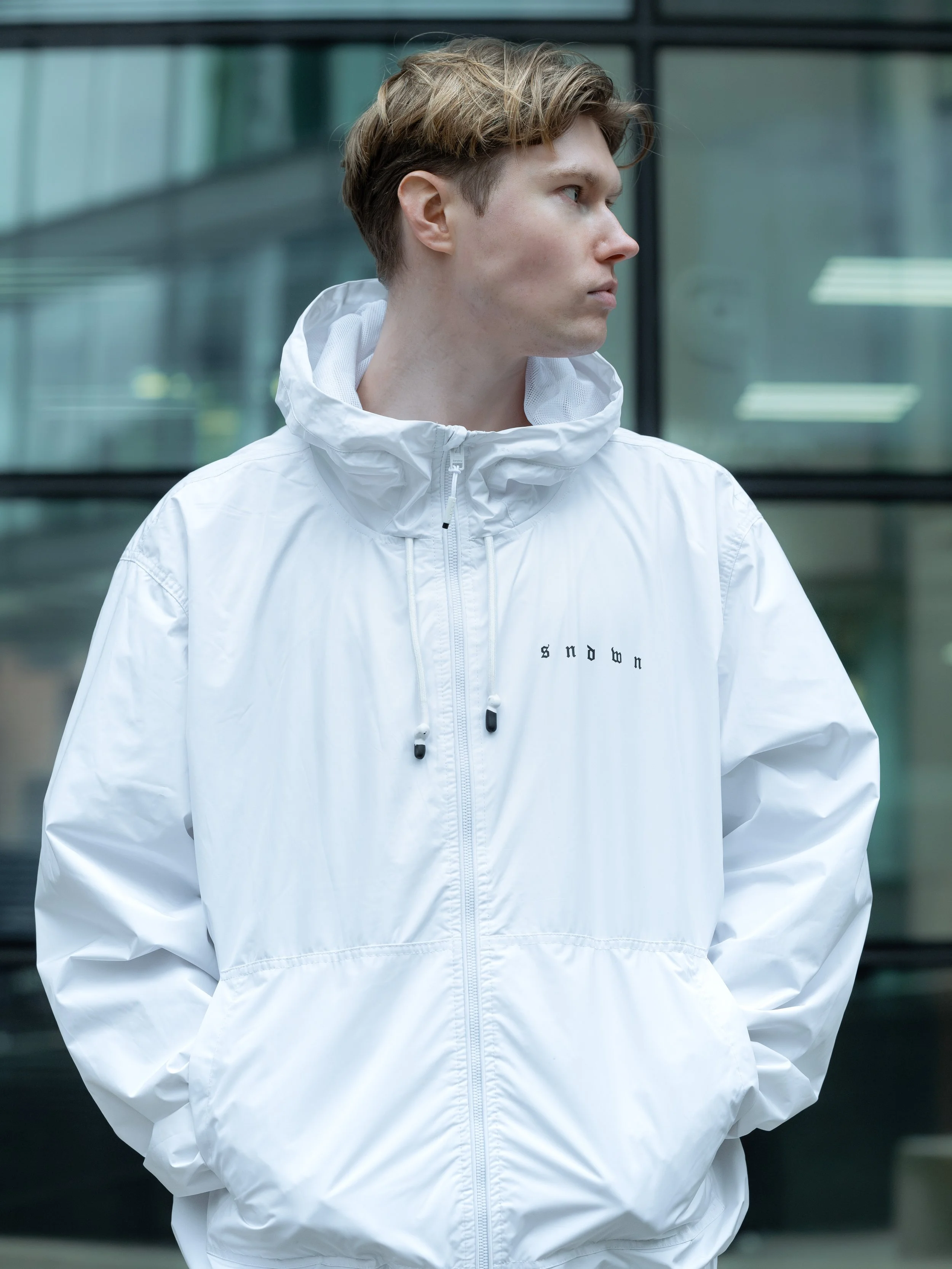 Young man with short, light brown hair wearing a white windbreaker jacket with a high collar, standing outdoors near a modern glass building, looking to the side.