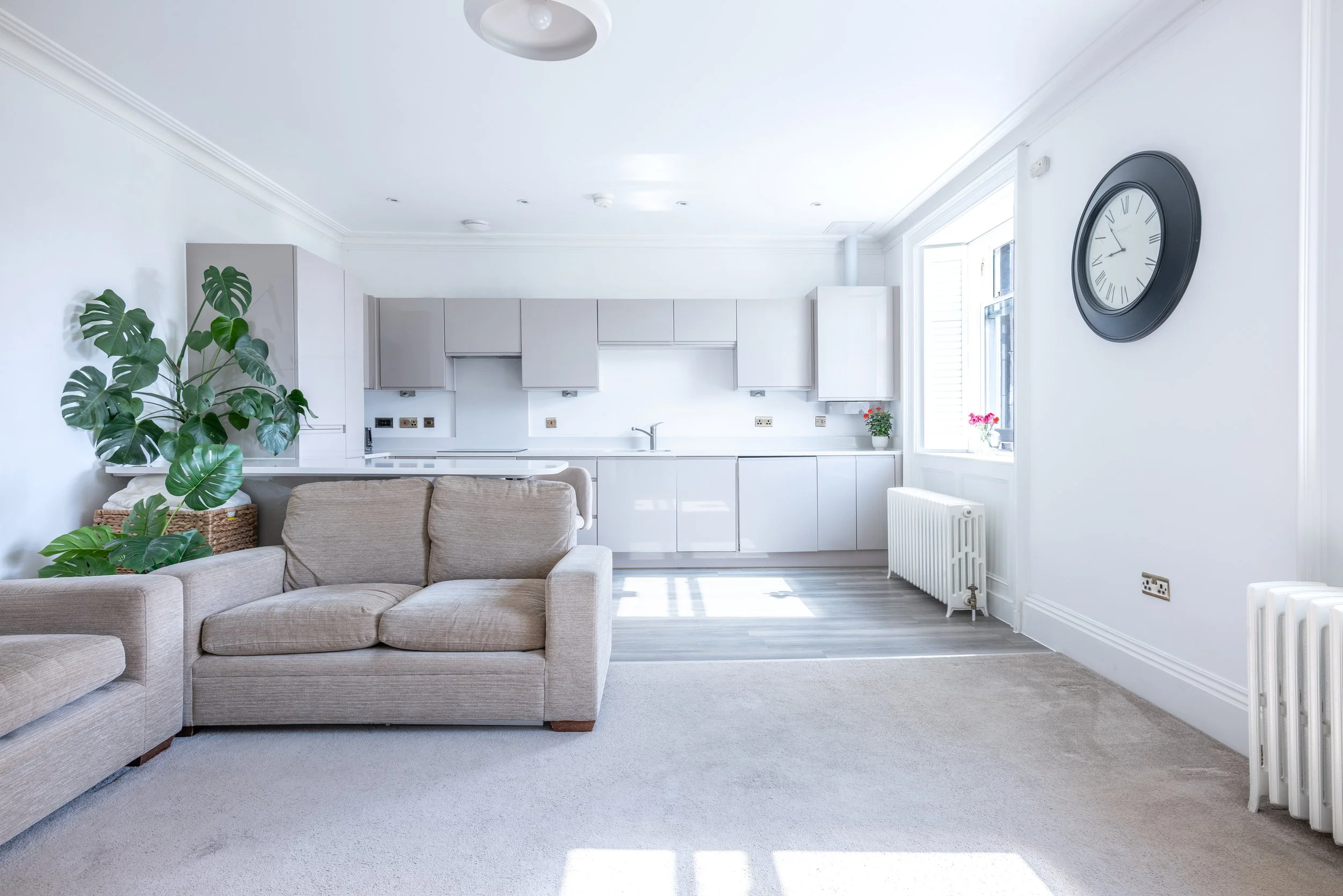 Bright open-plan living area with a beige sofa, a houseplant, and a white kitchen with gray cabinets, a large window with pink flowers, and a black wall clock.