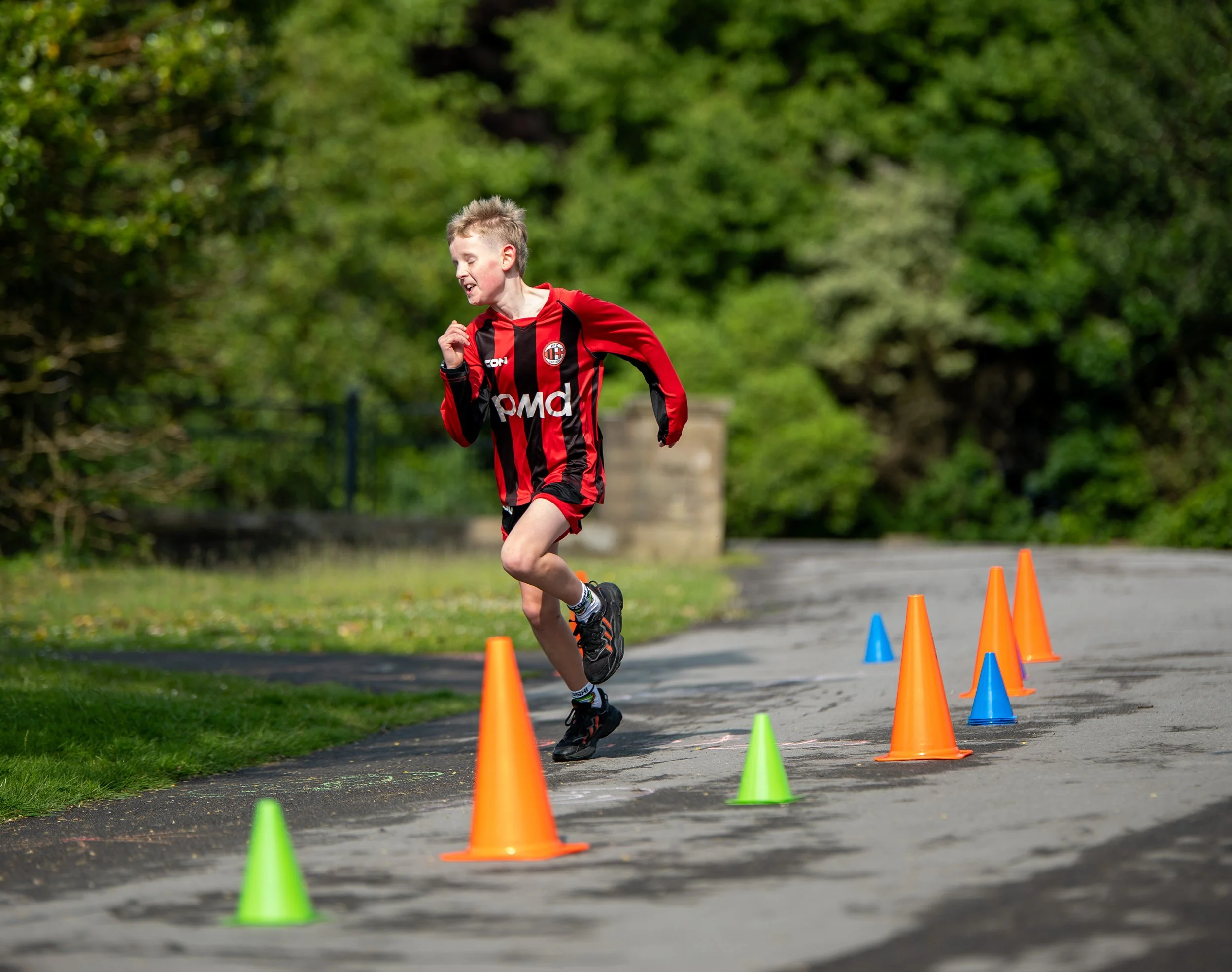 A young boy running on a paved pathway lined with colorful traffic cones during an outdoor activity on a sunny day with green trees in the background.