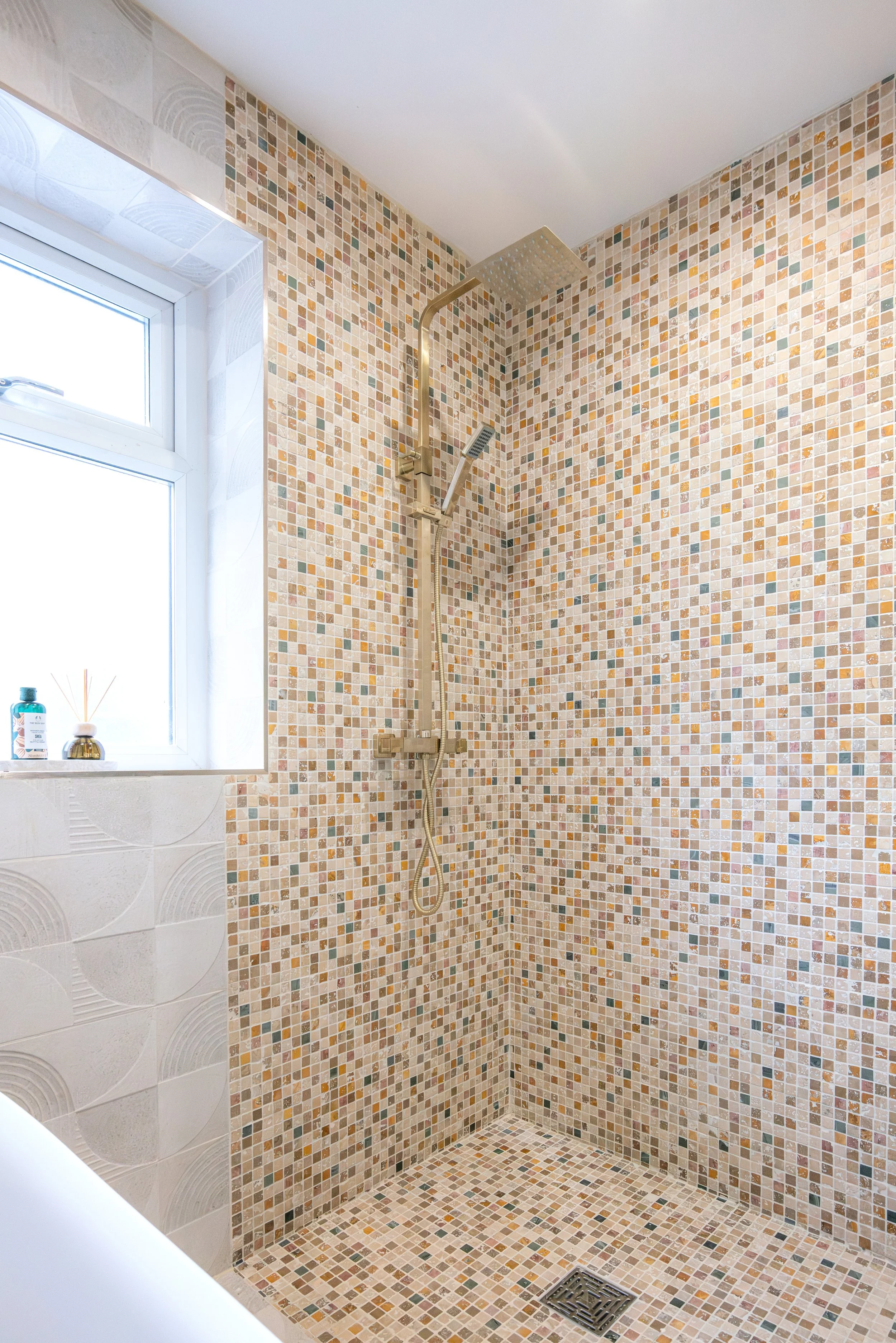 A walk-in shower with beige, brown, and tan mosaic tiles, a square rainfall showerhead, handheld showerhead, and a window with a bottle and reed diffuser on the sill.