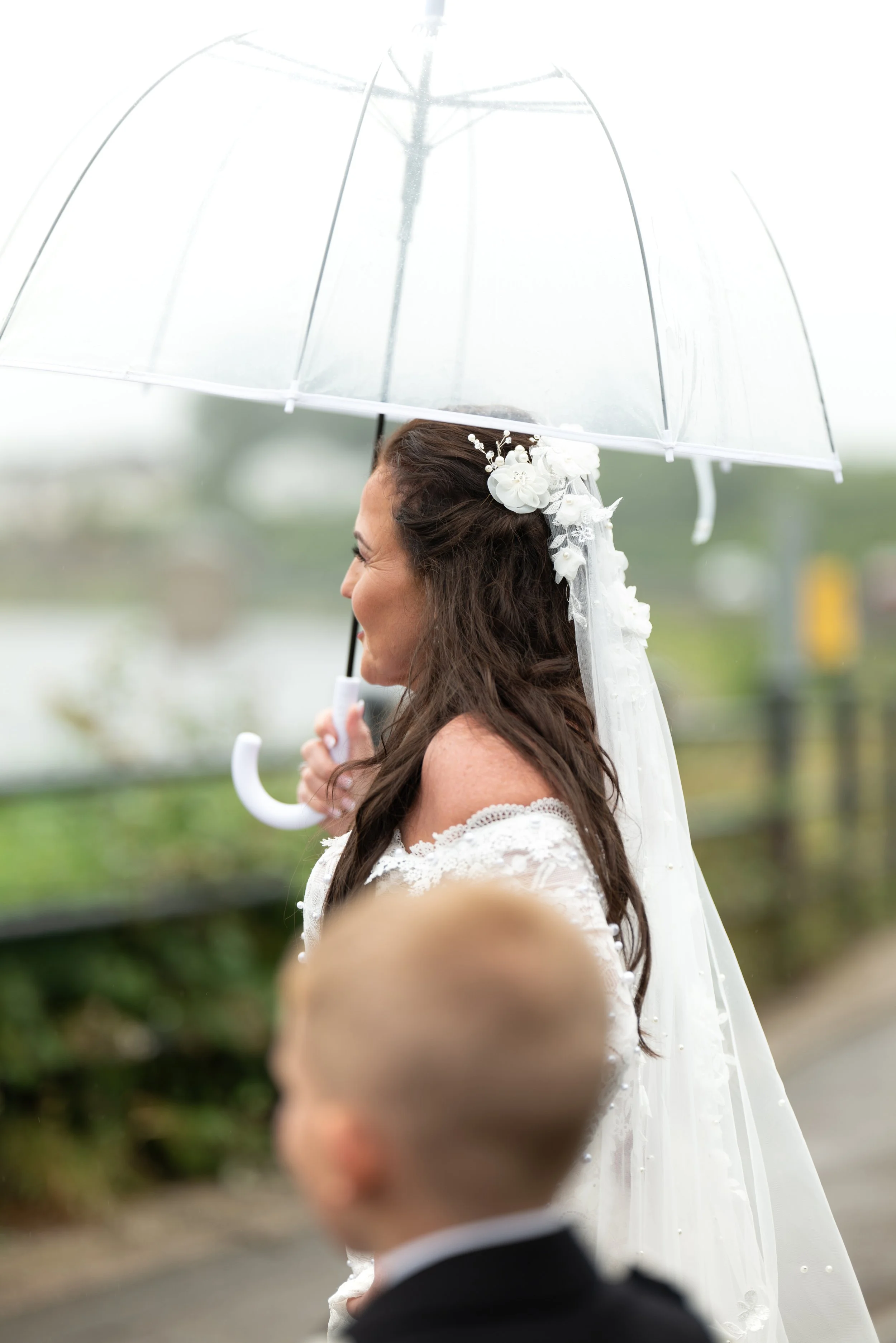 A woman wearing a wedding dress holding a transparent umbrella with a bride in the background, and a little boy in the foreground.
