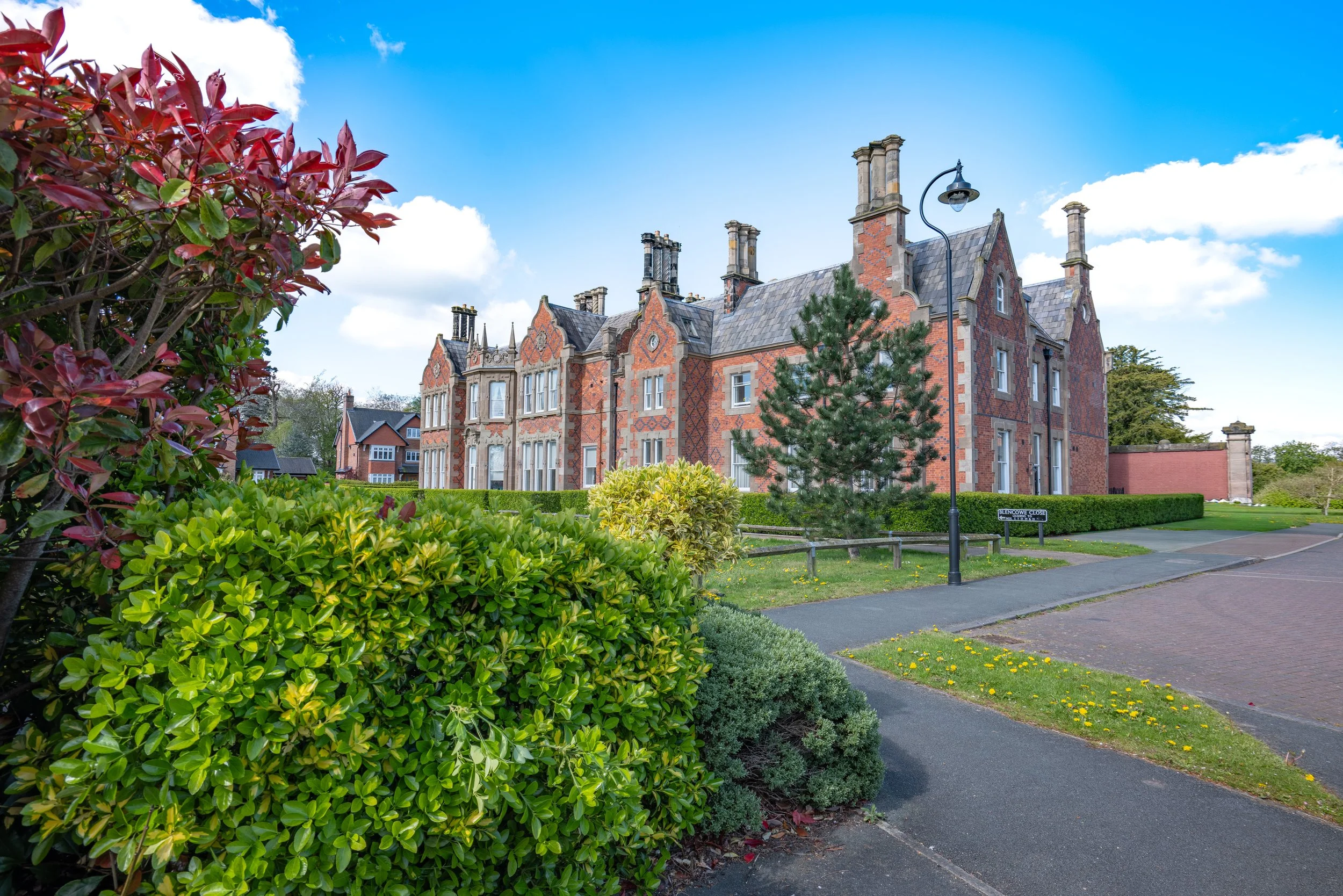 A historic brick building with towers and ornate architectural details, surrounded by a well-manicured garden with bushes, trees, and a sidewalk, under a bright blue sky with some clouds.