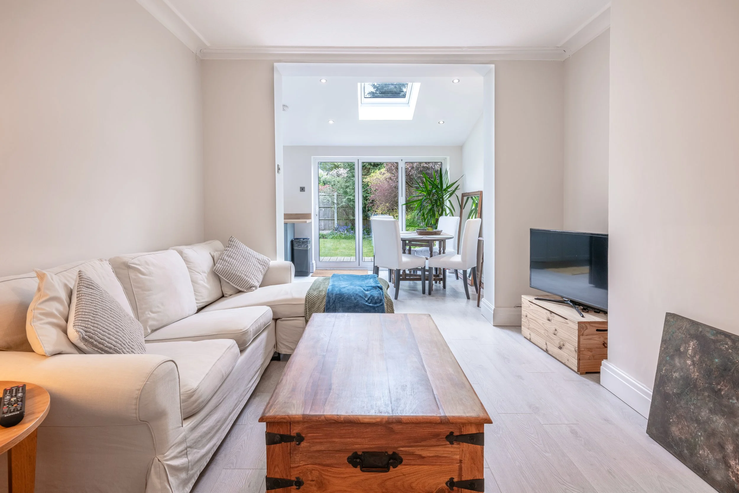 Living room with cream sofa, wooden coffee table, flat-screen TV on a wooden stand, dining area with round table and white chairs, large window and sliding door leading to a garden, skylight in ceiling, potted plant near the window.