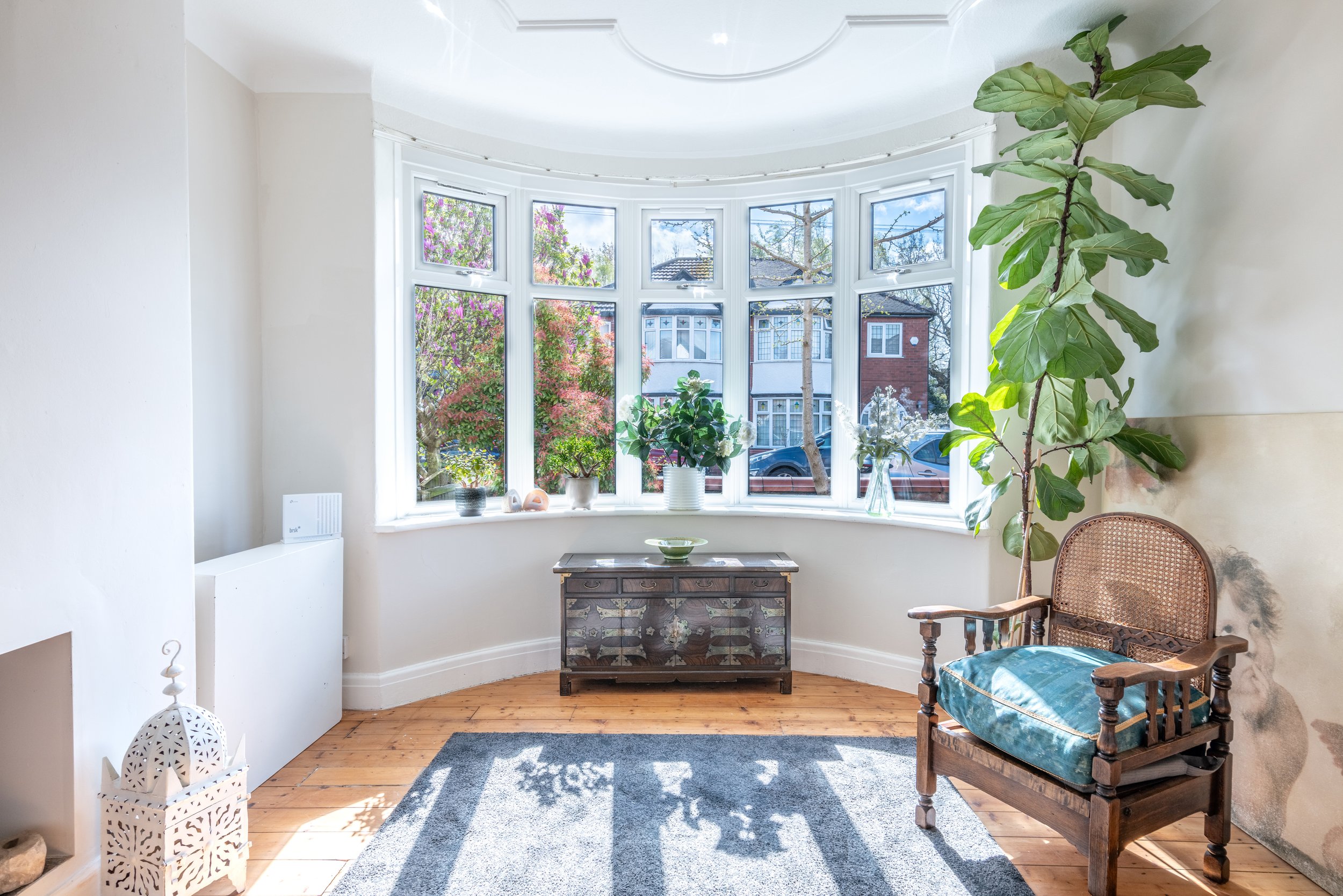 Bright living room with a large bay window, potted plants, a wooden chair with a blue cushion, a gray rug, and a small vintage cabinet. Sunlight streams through the window, casting shadows on the wooden floor.