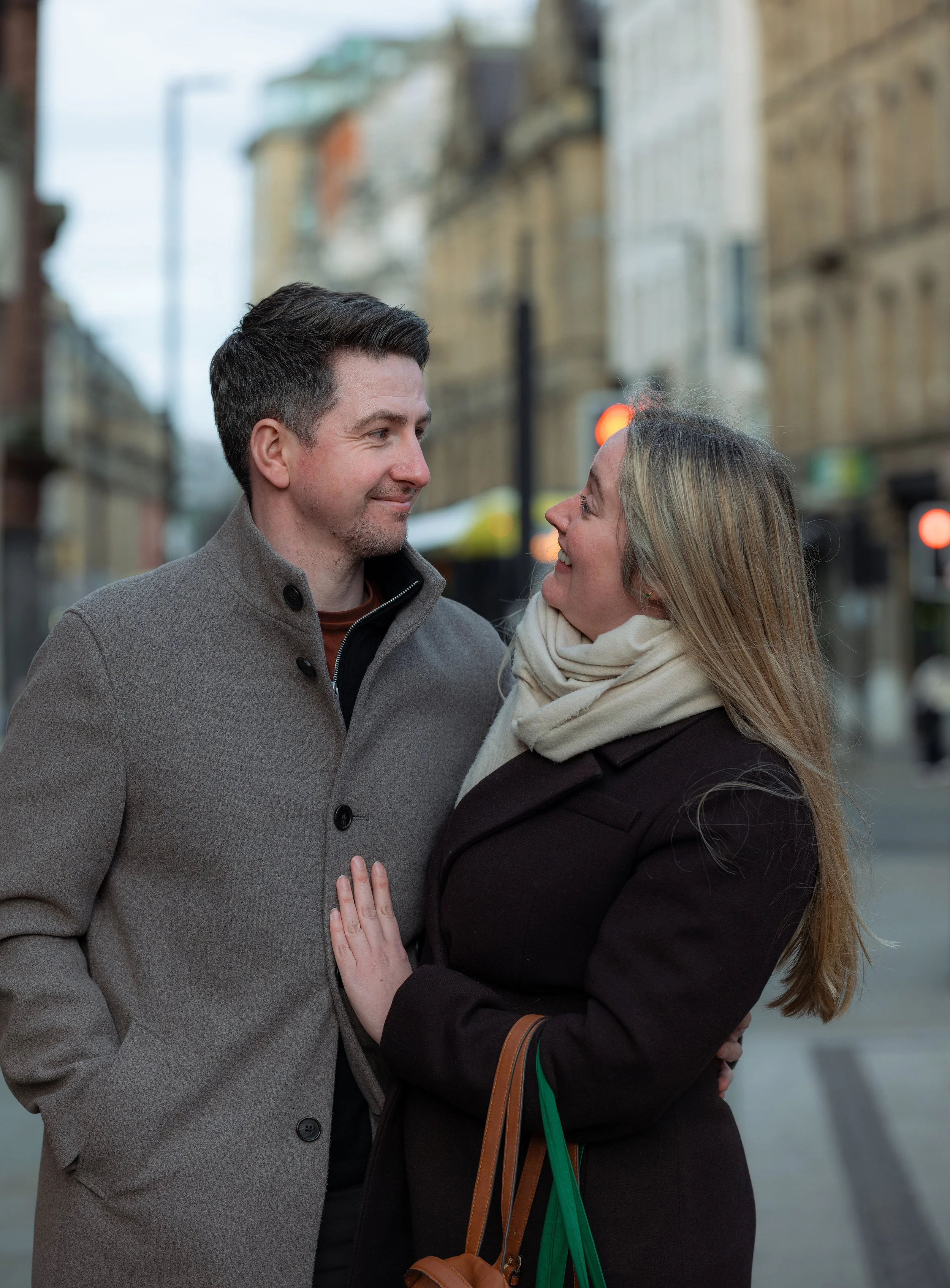 A man and woman standing close together on a city street, smiling and looking into each other's eyes, dressed in coats and scarves.