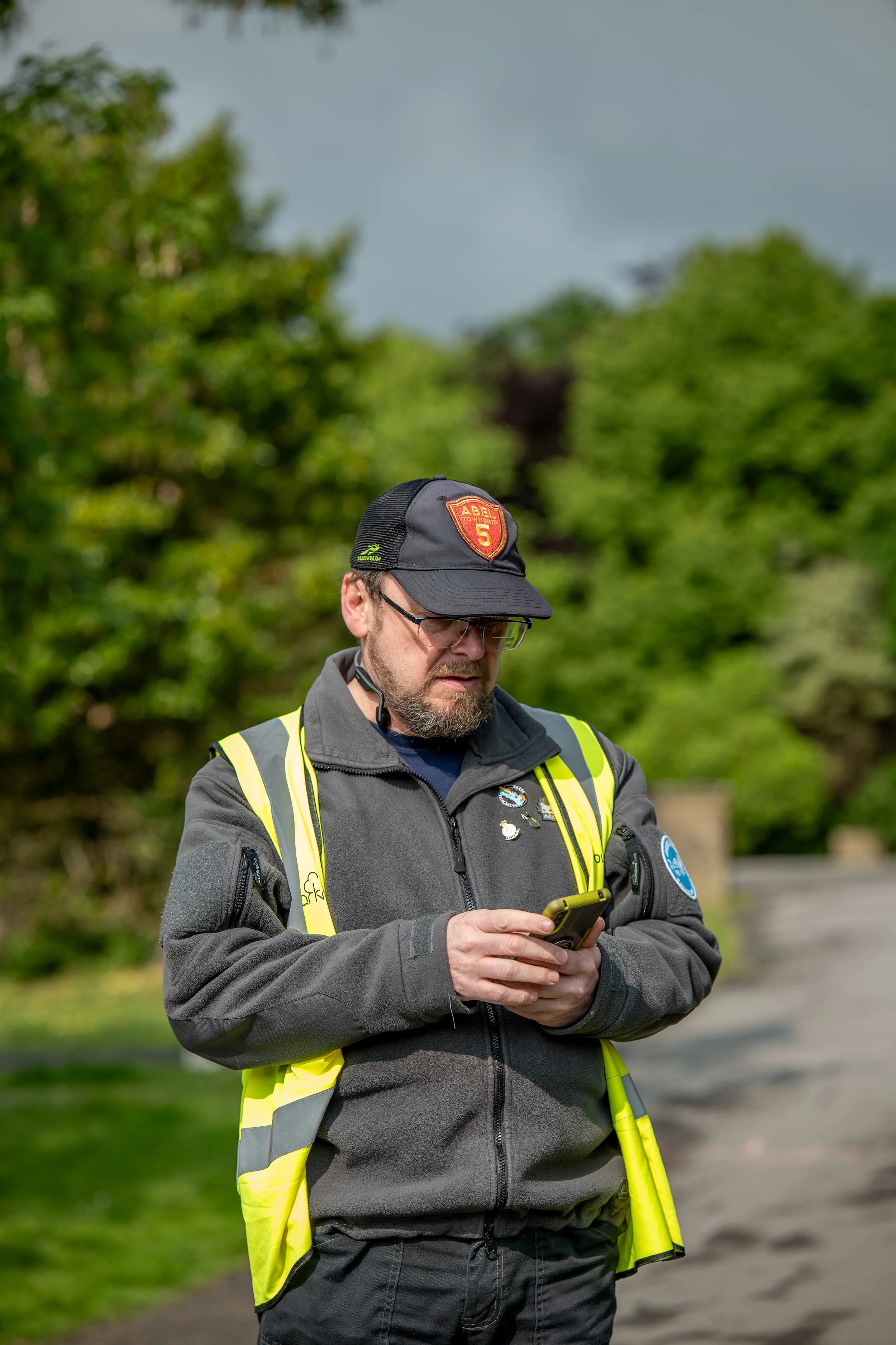A man with a beard and glasses looking at his phone outdoors, wearing a gray jacket, a yellow safety vest, and a black cap with a red and yellow patch, with green trees and a cloudy sky in the background.