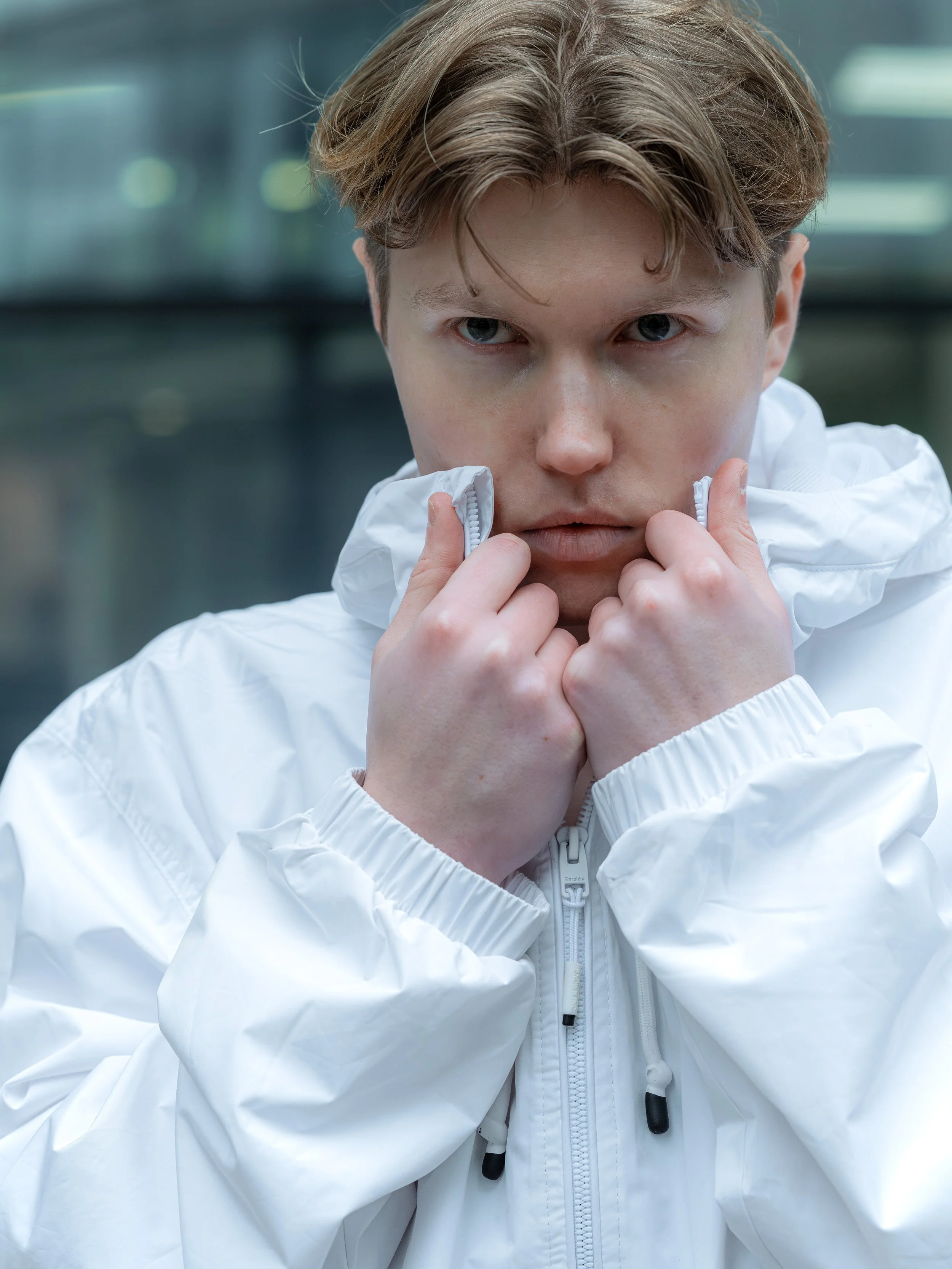 A young man with blonde hair in a white jacket is looking directly at the camera, holding the collar of his jacket with both hands.