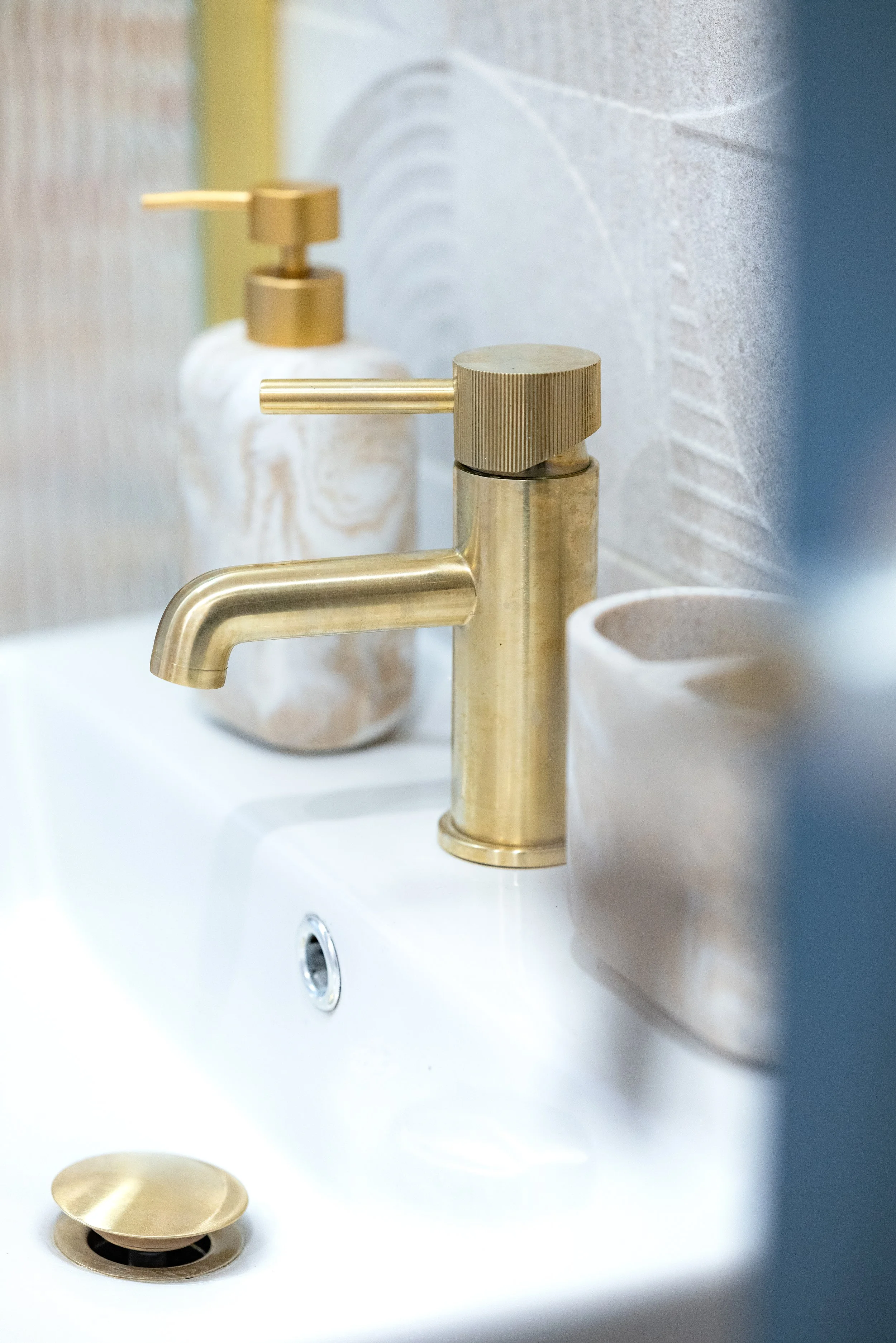 Close-up of a modern gold-colored bathroom faucet mounted on a white sink, with a soap dispenser and toothbrush holder visible in the background.