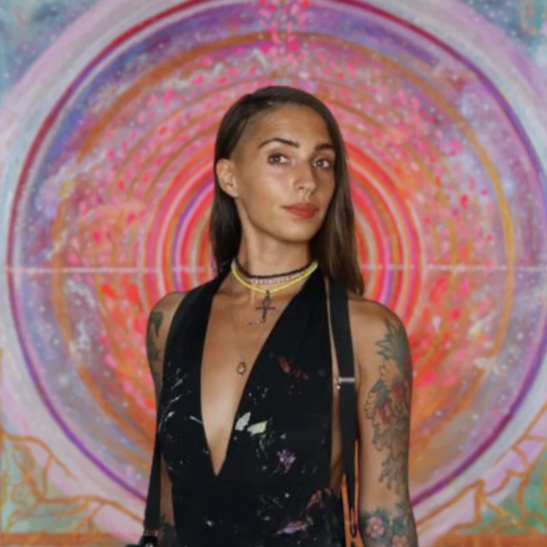 A woman with dark brown hair and tattoos on her arms is standing in front of a vibrant, colorful background with concentric circles and pink, purple, and orange hues.