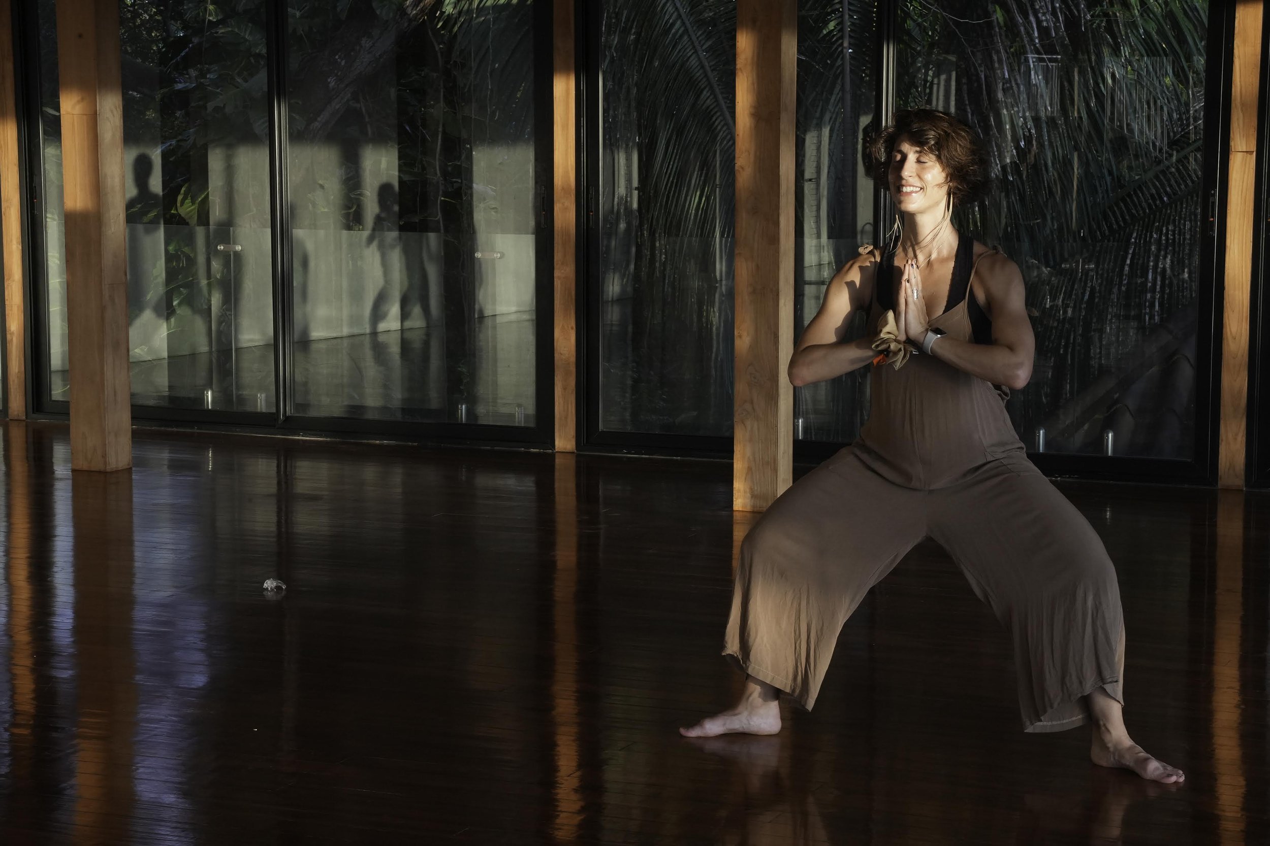 A woman practicing yoga in a spacious room with large windows and wooden floors, standing in a warrior pose with hands in prayer position.