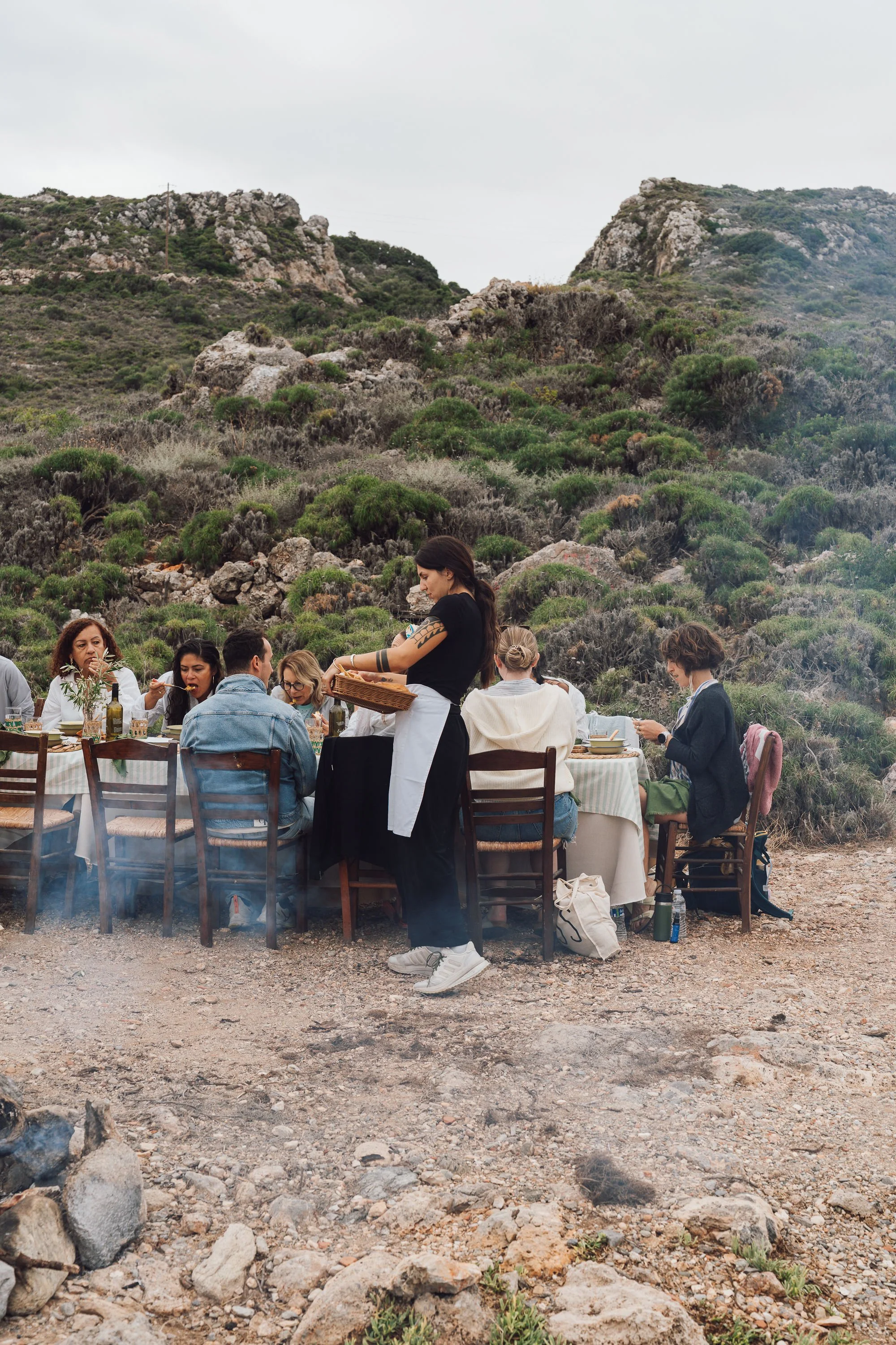 A group of people dining outdoors at a long table set on rocky terrain with a mountainous landscape in the background, while a server in black and white attire serves them.