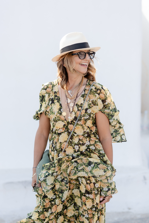 Woman wearing a floral dress, hat, glasses, and layered necklaces, smiling and looking to her right.