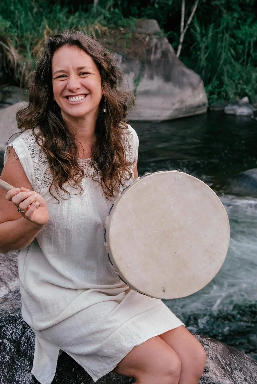 A smiling woman with long wavy brown hair sitting on a rock by a stream, holding a drum with a drumstick in her hand, surrounded by green foliage.