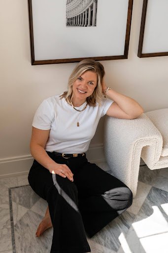 Woman sitting on the floor, smiling, with artwork on the wall behind her.