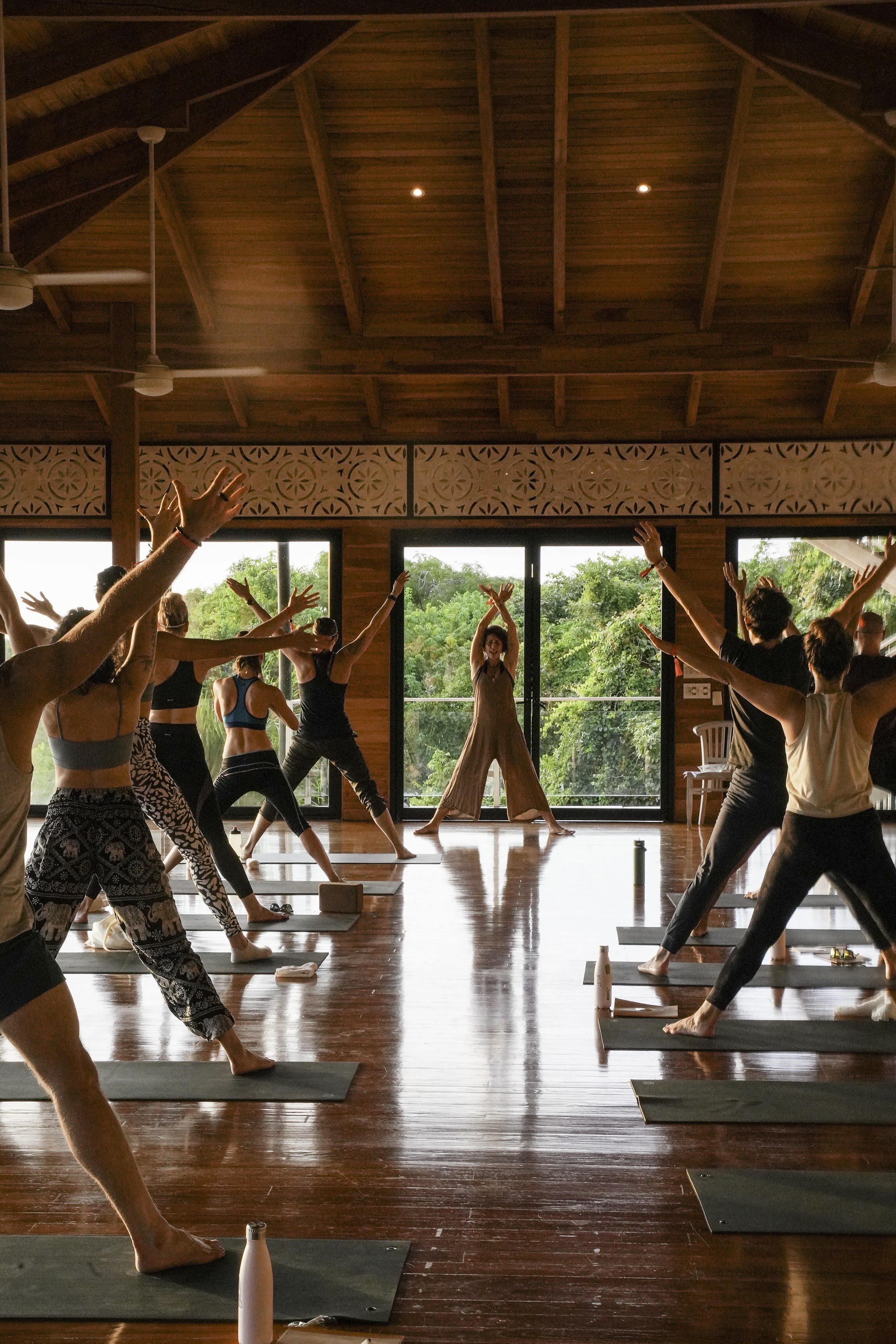 Indoor yoga class with students in a spacious wooden-floored room, raising arms, led by instructor at front, large windows showing greenery outside.