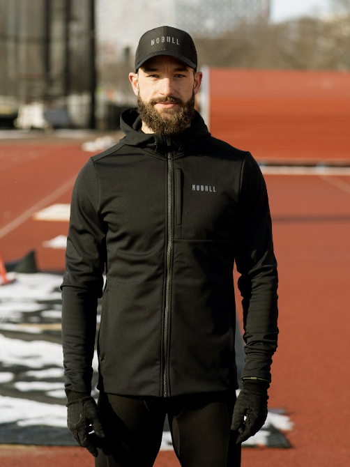 Man standing outdoors on a running track, wearing a black Nike hooded jacket, matching cap, black gloves, and sportswear, with snow patches and sports equipment in the background.