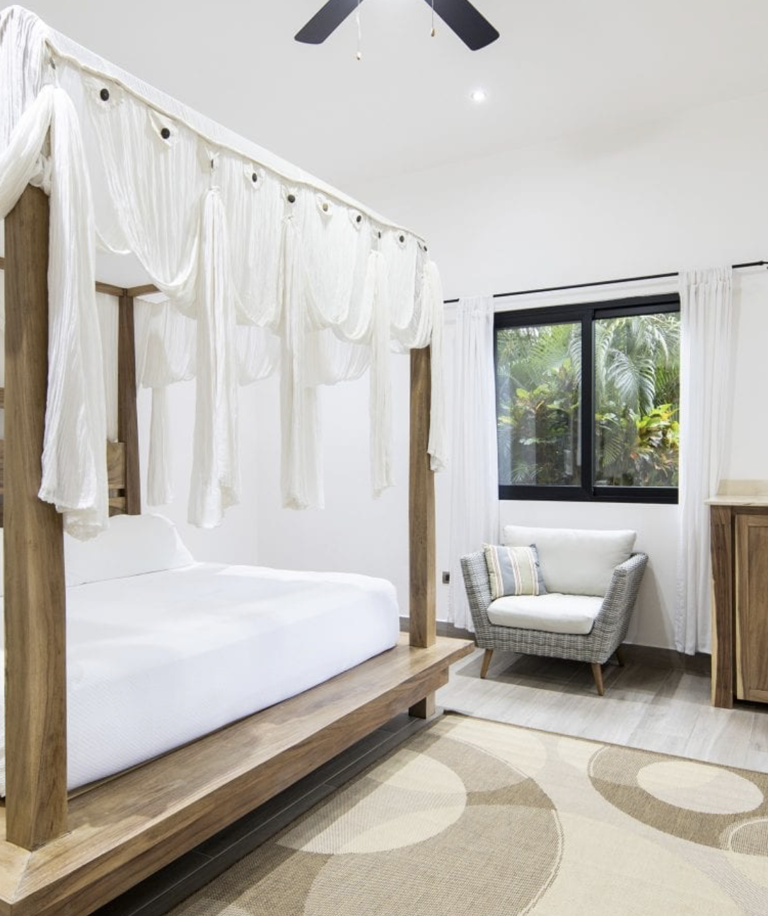 A bedroom with a wooden four-poster bed with white drapes, a window with white curtains, a wicker armchair with a striped pillow, and tropical greenery outside the window.