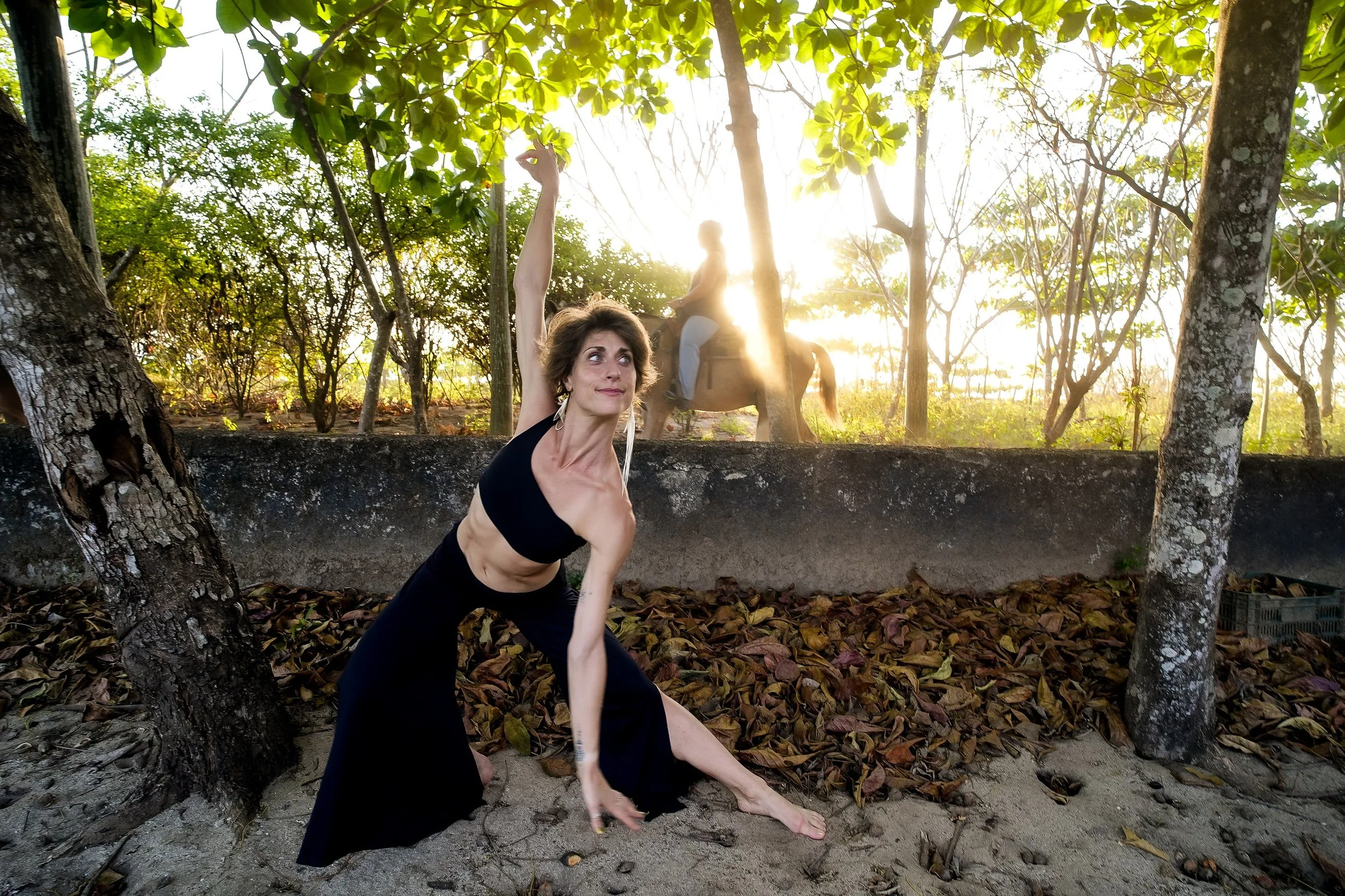 A woman practicing yoga outdoors on sandy ground next to trees, with a woman riding a horse in the background during sunset.