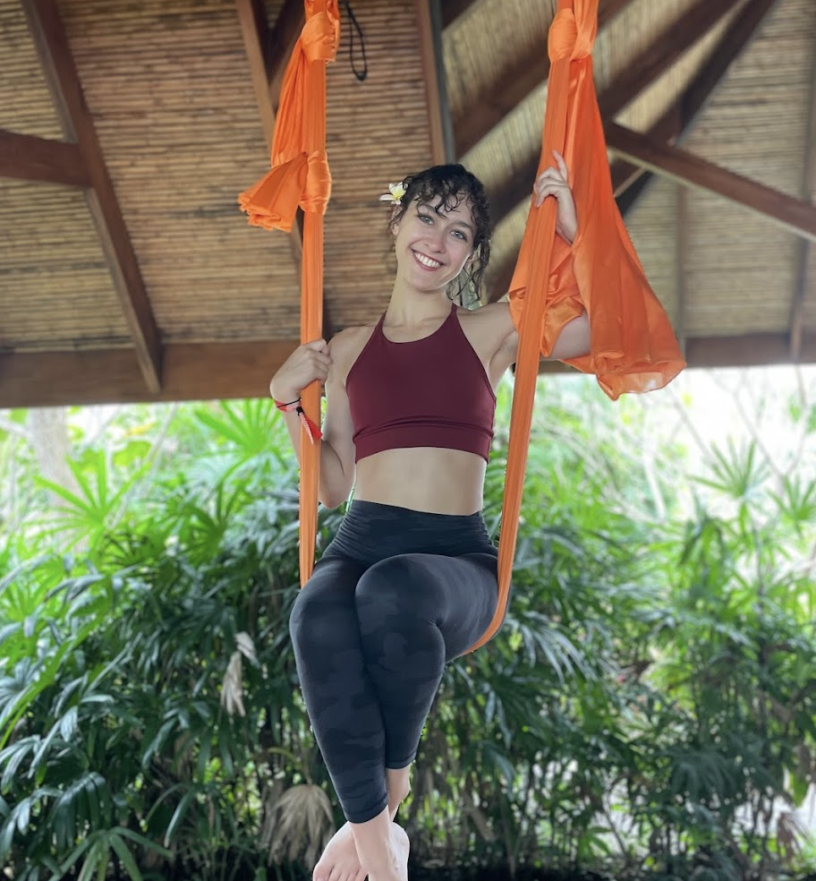 A woman smiling on orange aerial yoga swing indoors with green plants and wooden ceiling in the background.
