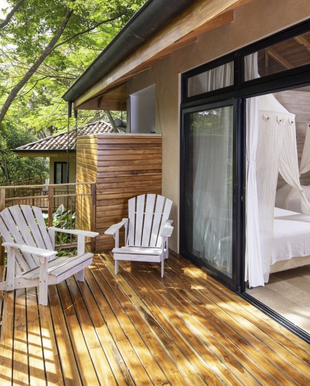 A wooden deck with two white Adirondack chairs, surrounded by green trees, attached to a house with sliding glass doors and white curtains inside.