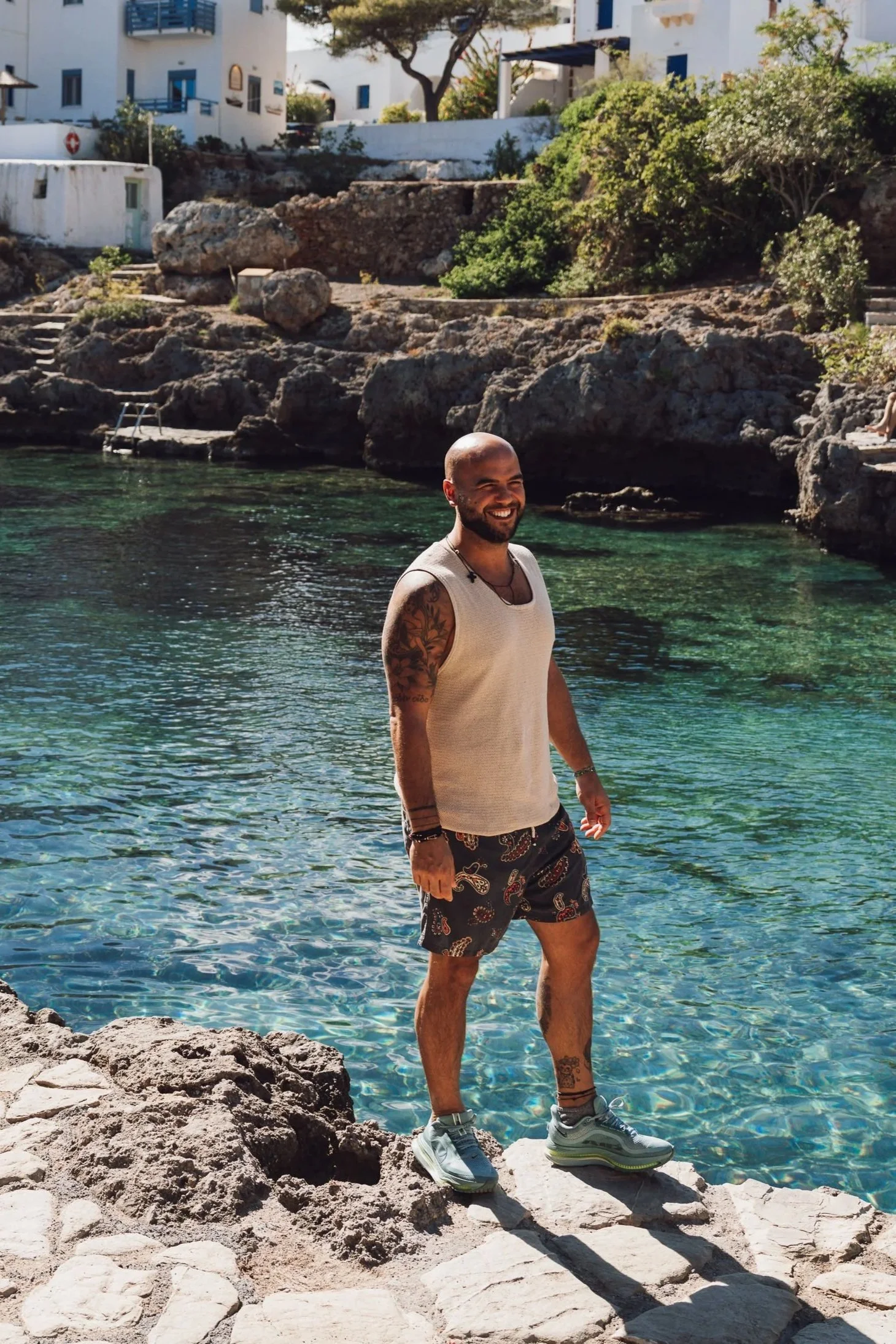 A smiling man with tattoos, wearing a sleeveless shirt, patterned shorts, and sneakers, standing on rocks by clear water with a rocky shoreline and white buildings in the background.