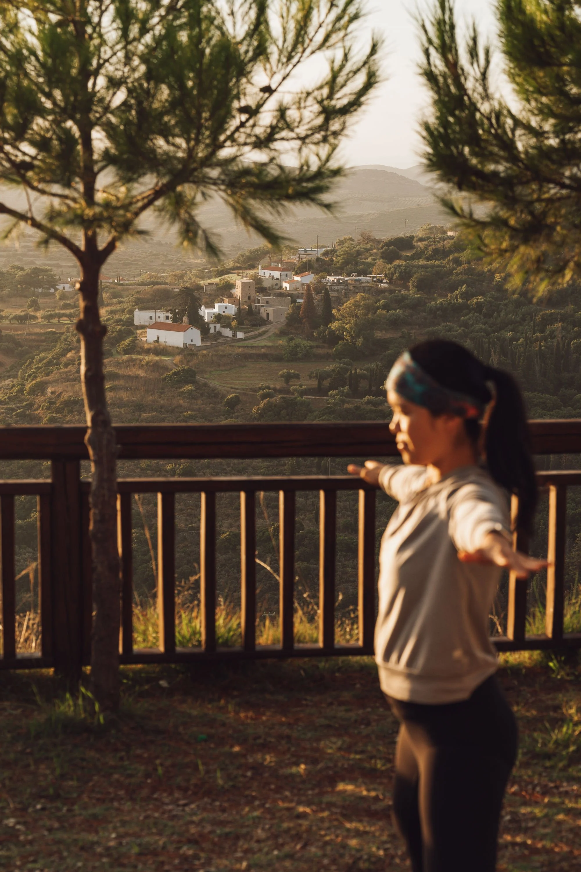 A woman with dark hair in a ponytail and a blue headband stands outdoors on a wooden deck, with arms outstretched and eyes closed, facing a scenic landscape of rolling hills, trees, and white houses at sunset.