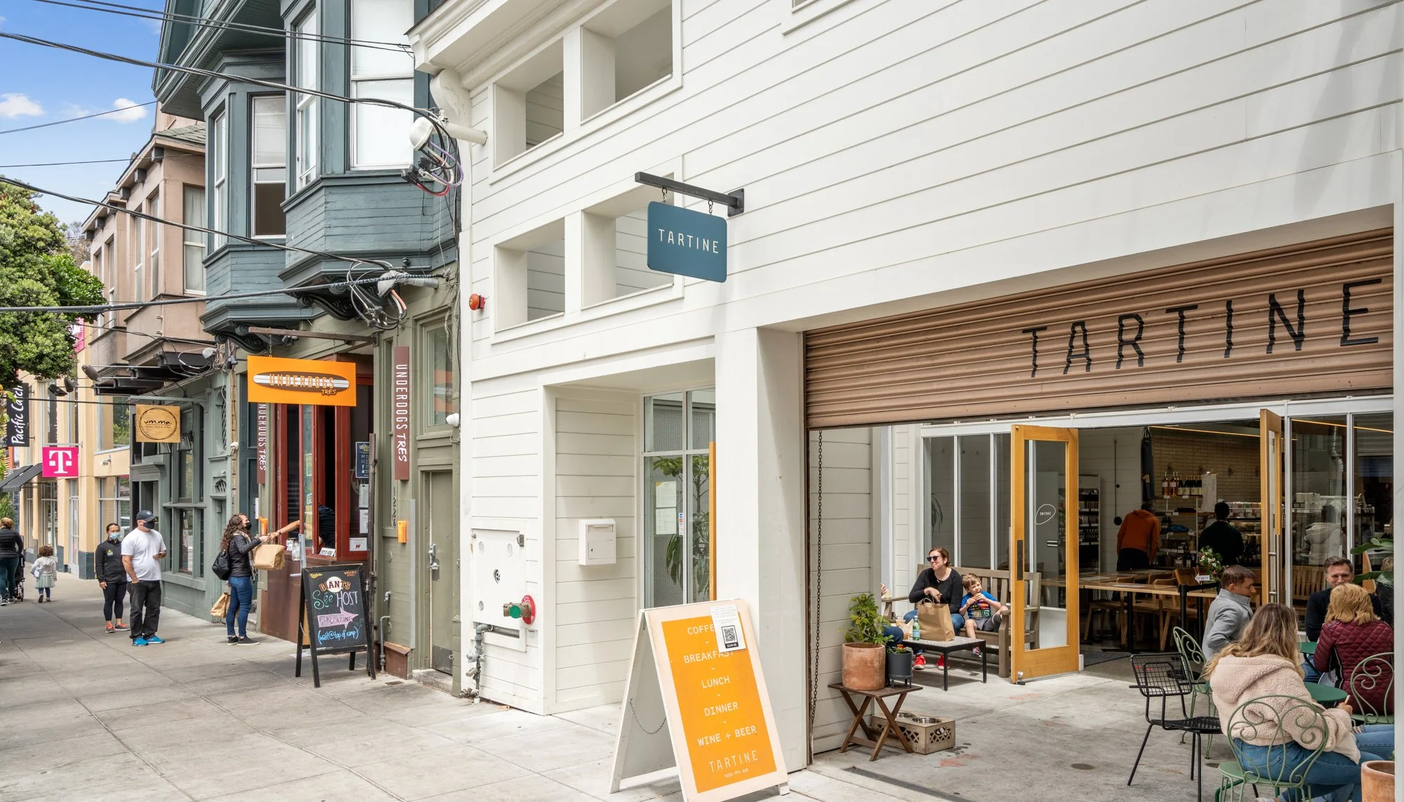 Street scene with people dining outside a cafe named Tartine, with others walking along the sidewalk, storefronts, and a sign advertising coffee, breakfast, lunch, dinner, and wine and beer.