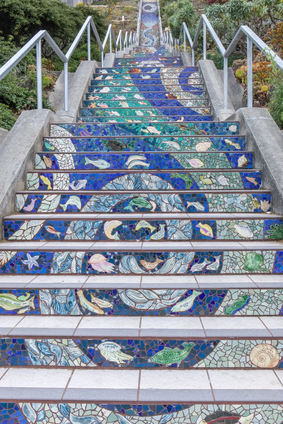 Colorful mosaic tiled staircase depicting ocean and marine life, ascending through a garden with plants and trees on sides.