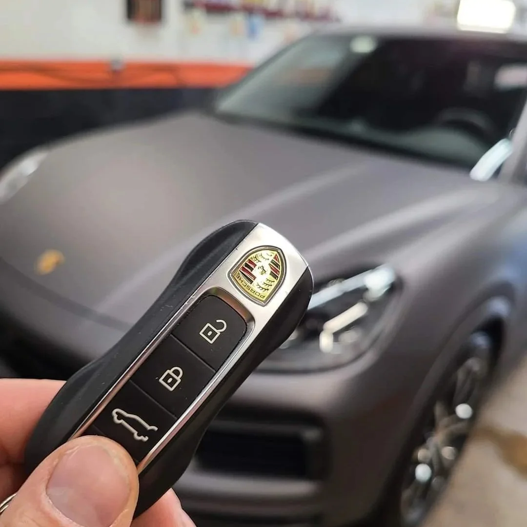 Close-up of a person's hand holding a Porsche key fob in front of a gray Porsche sports car.