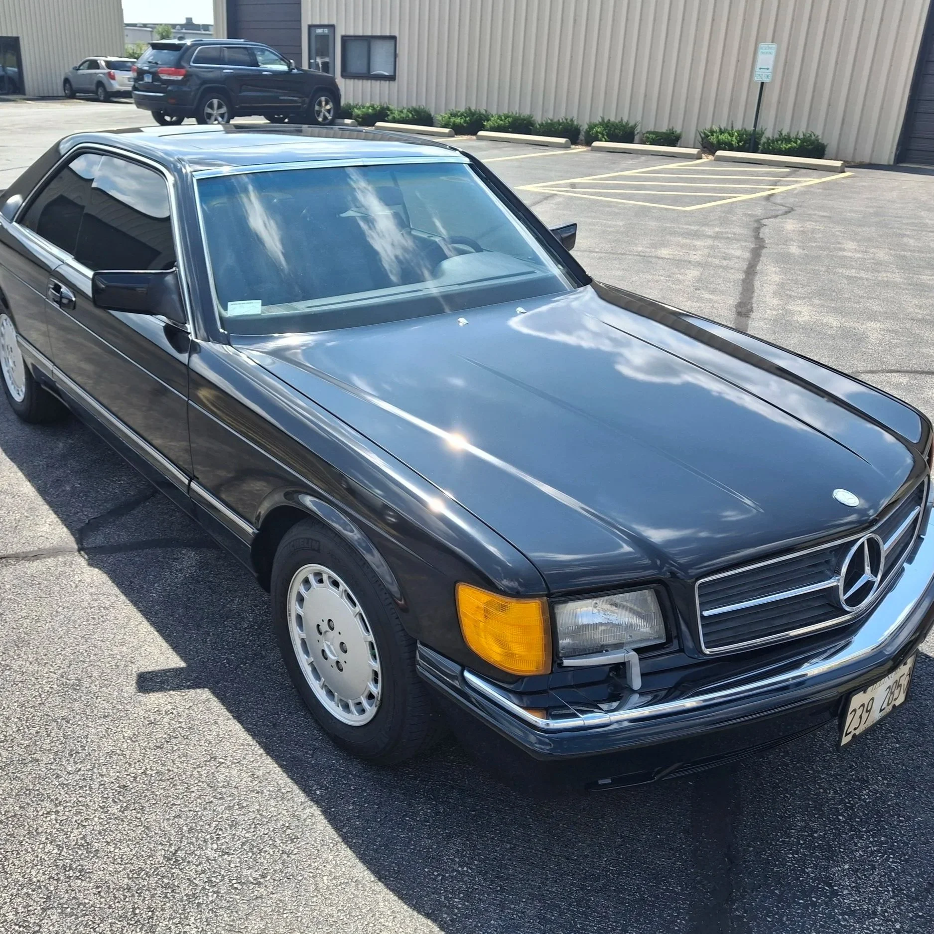 Black vintage Mercedes-Benz coupe parked in a lot, with other cars and a beige building in the background.