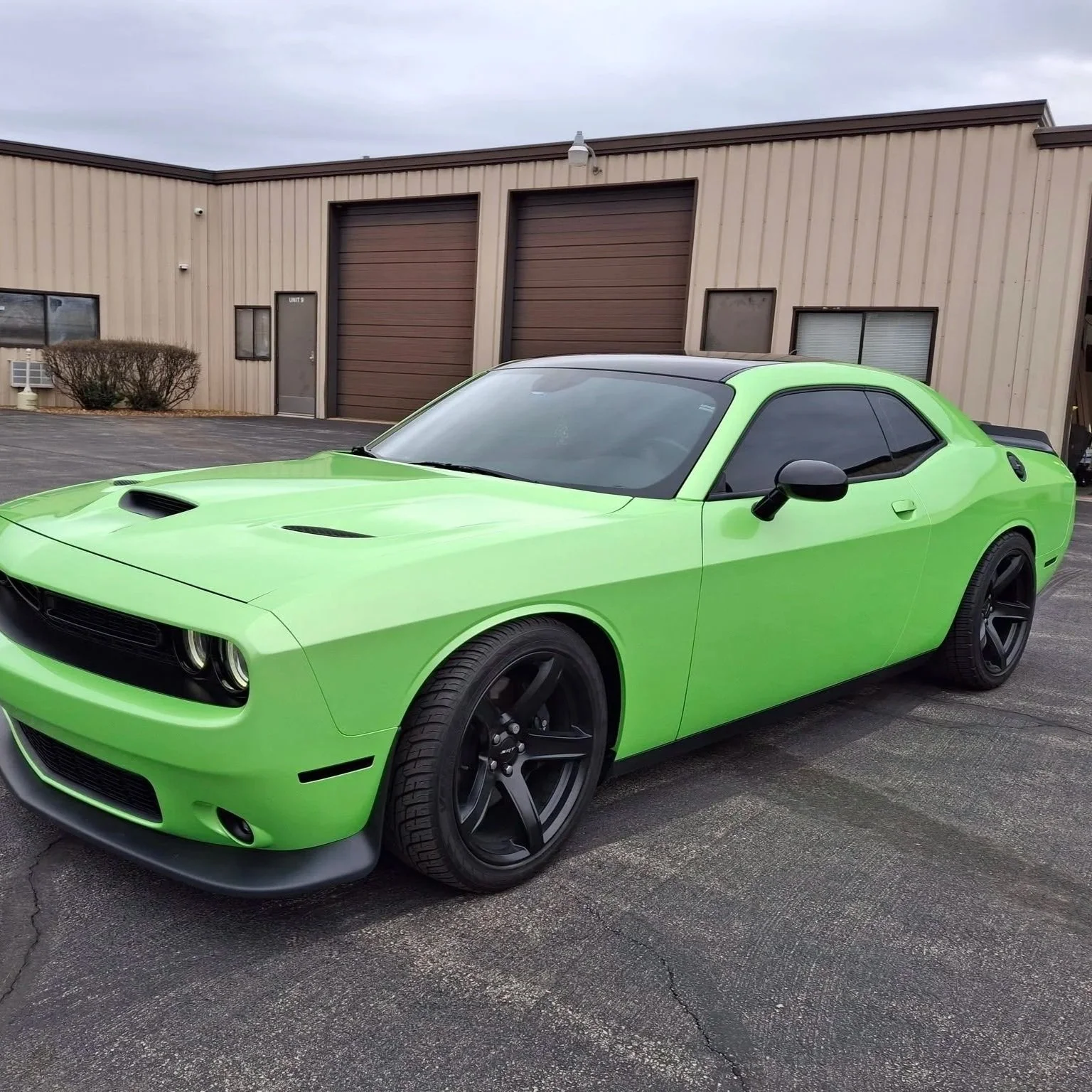 A bright green muscle car with black wheels parked outside a beige industrial building with brown garage doors.