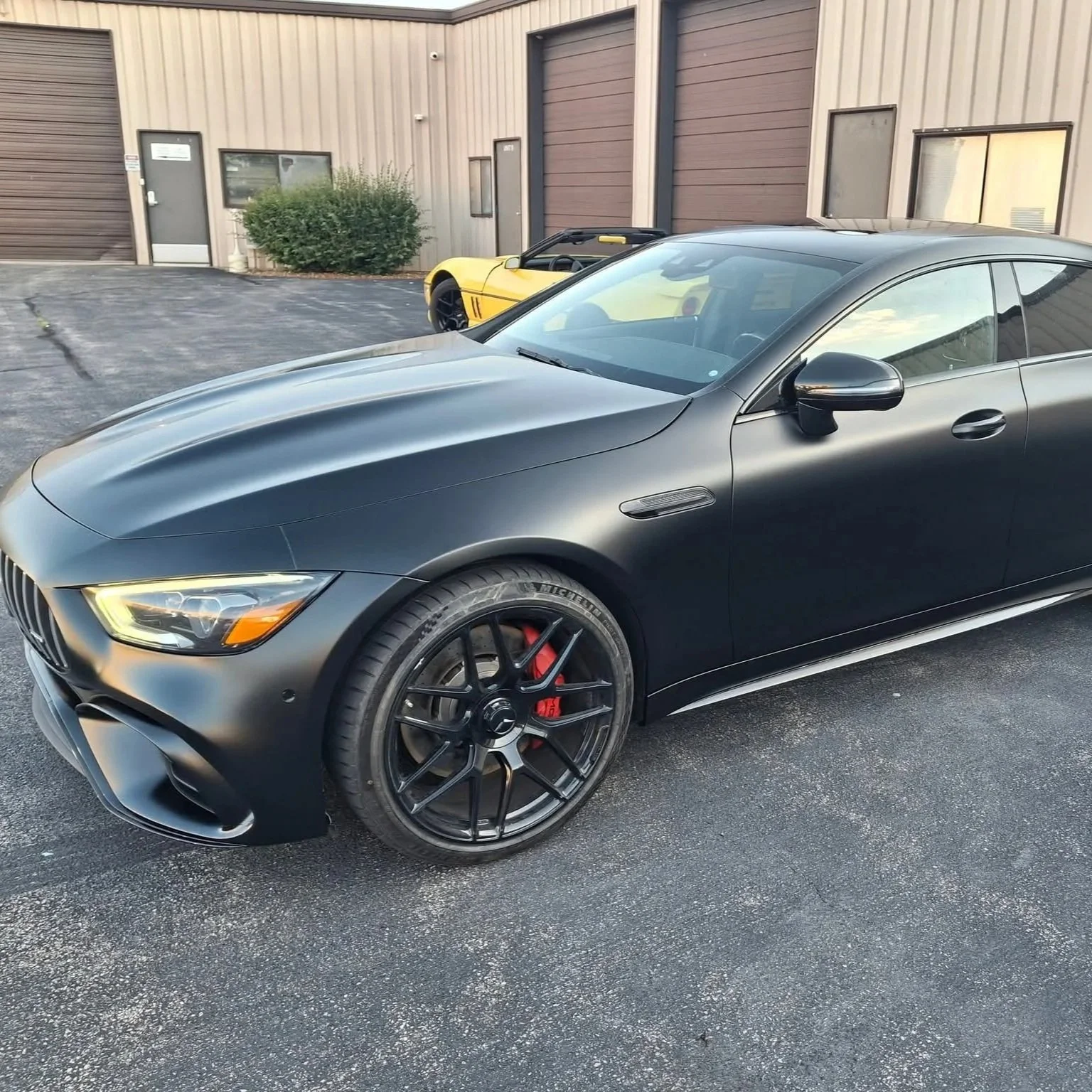Black luxury sports car parked in front of a beige industrial building with a yellow sports car in the background.