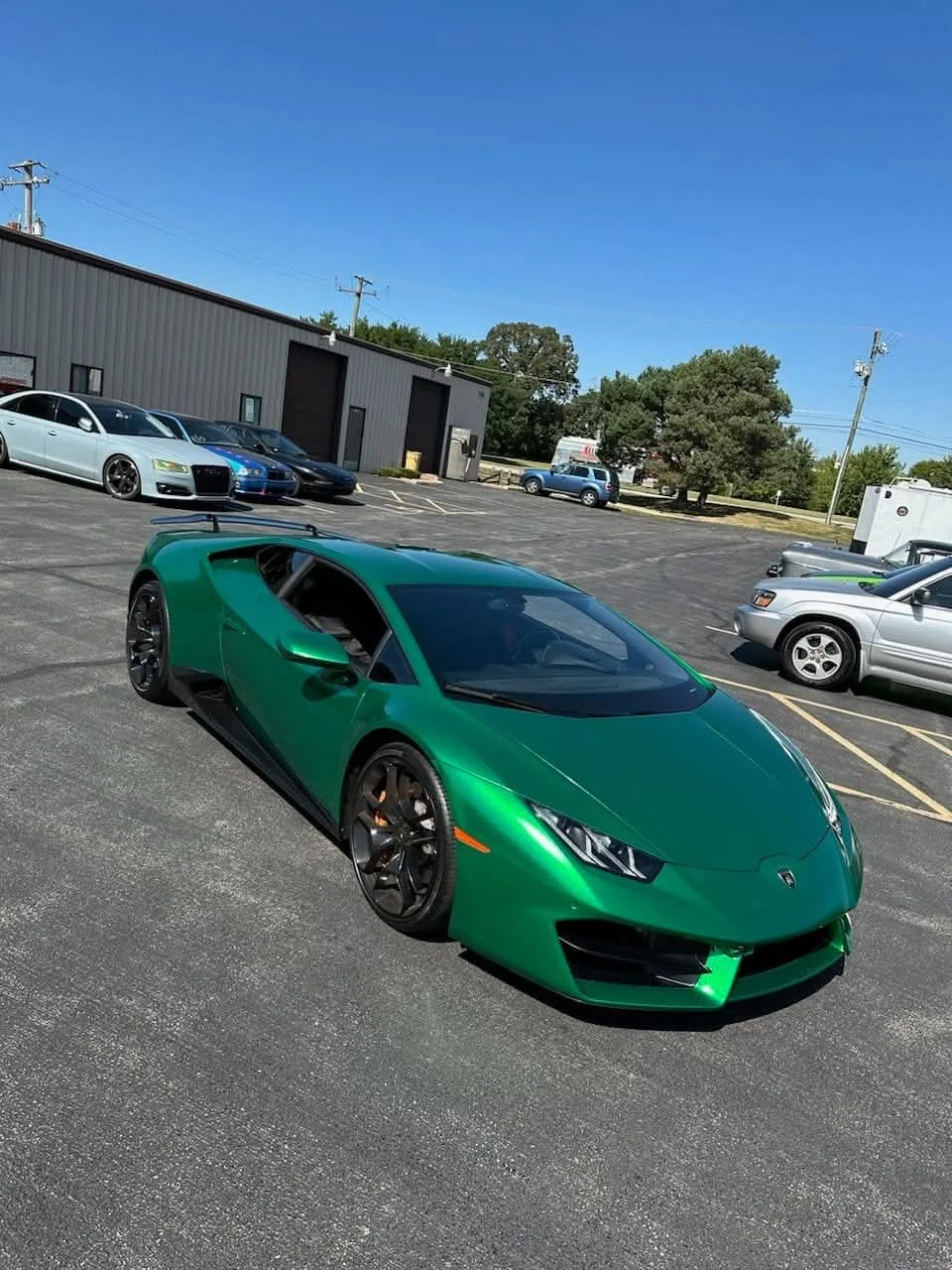 Green Lamborghini sports car parked in a lot with other vehicles and a gray industrial building in the background.