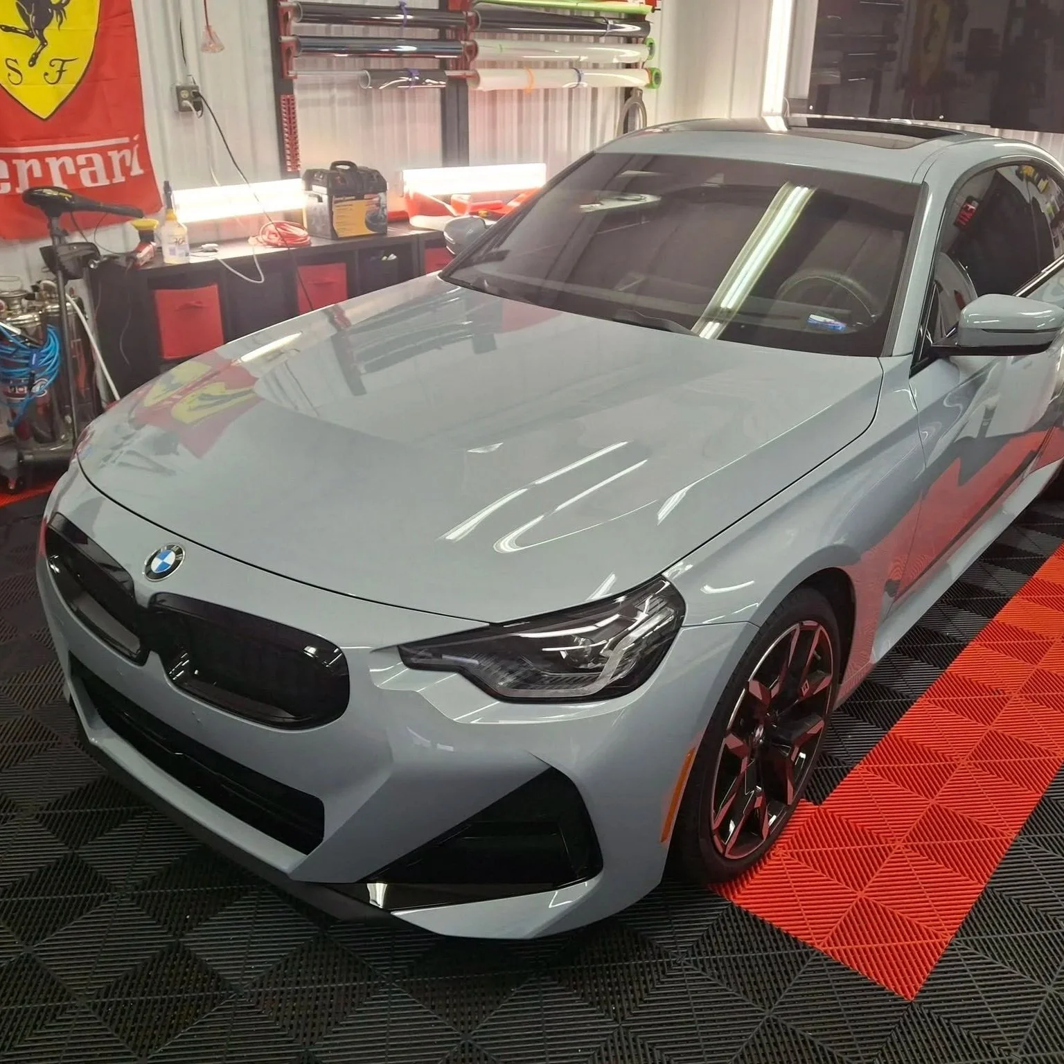 A silver BMW car parked in a garage or workshop with automotive tools and equipment visible in the background.
