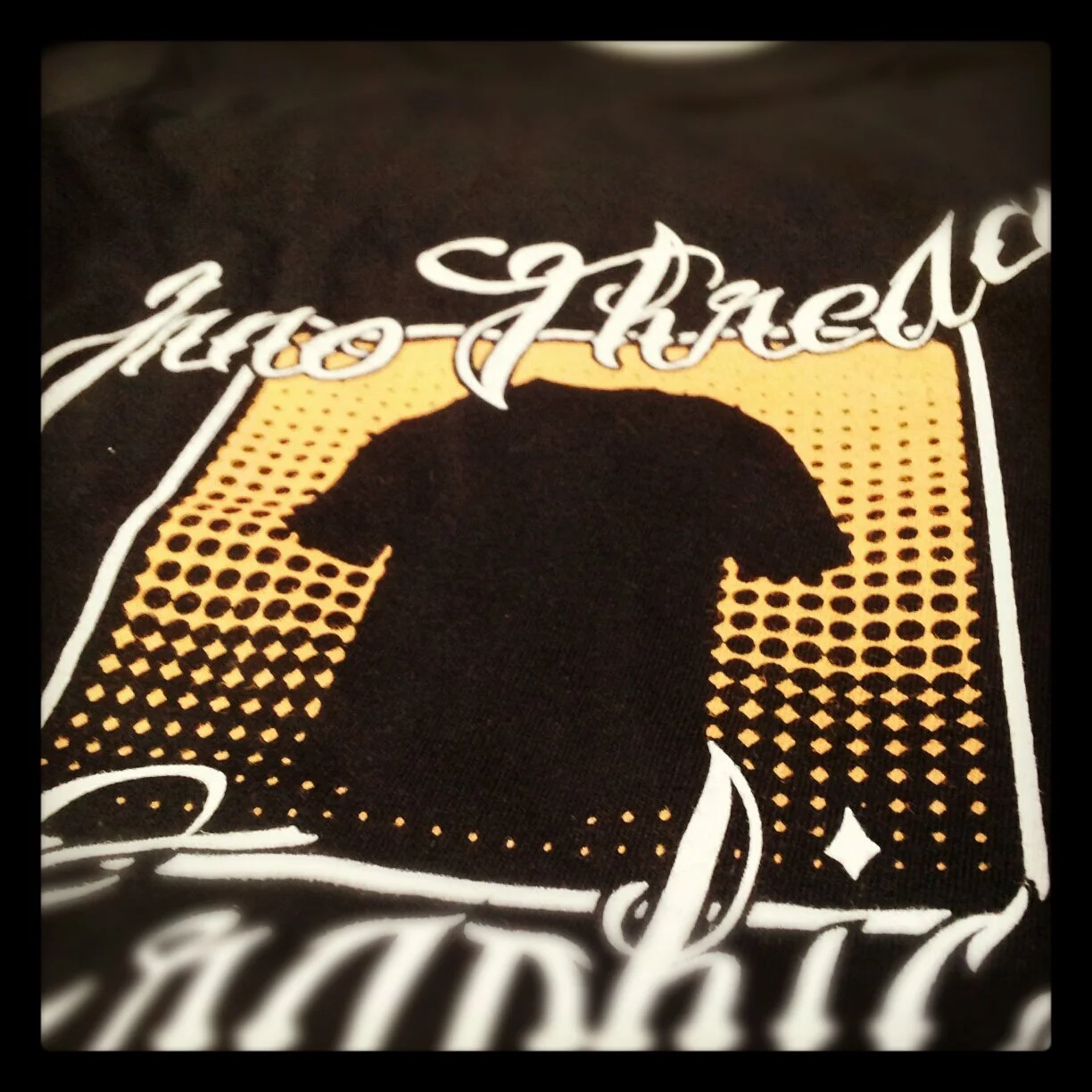 Close-up of a black T-shirt with orange and white graphic design, featuring a silhouette of a person's head in the center, surrounded by stylized text and dotted background patterns.
