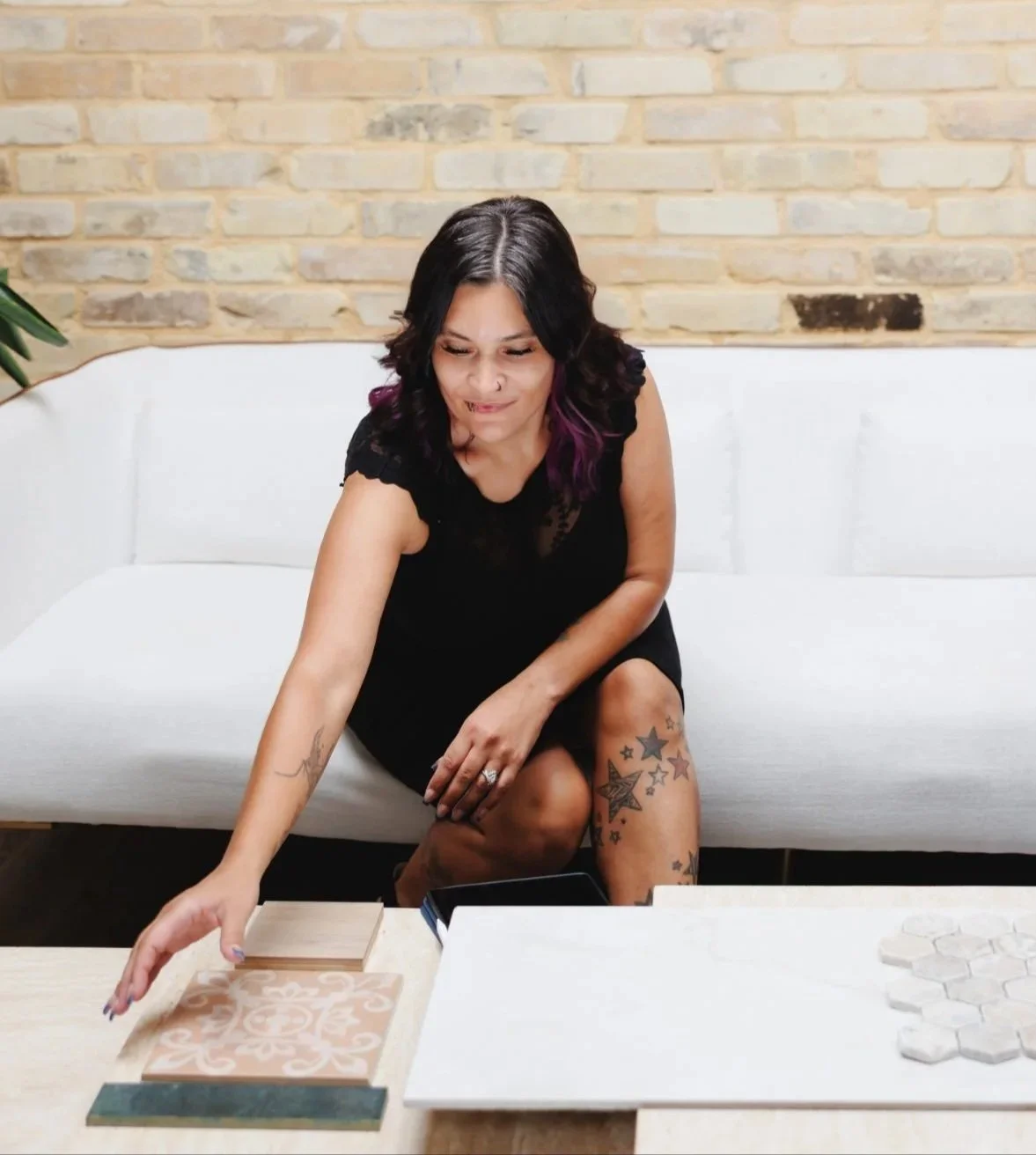 A woman with black hair and purple highlights sitting on a white couch in front of a brick wall, reaching for samples of tile or flooring on a table.