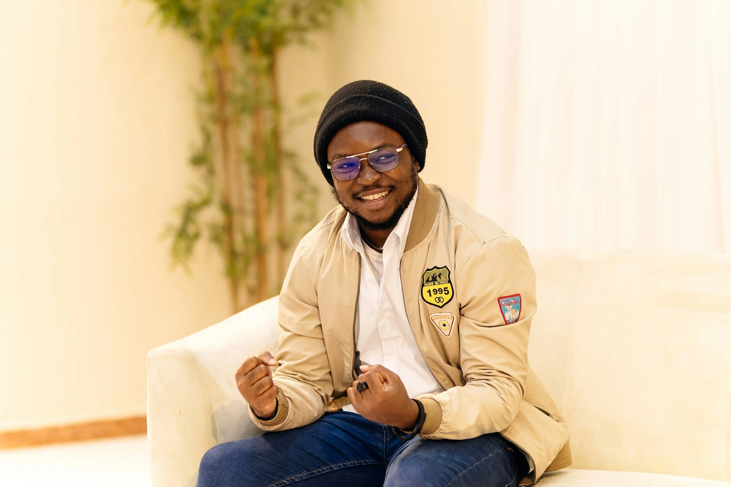 A smiling man wearing glasses, a black beanie, beige jacket with patches, white shirt, and dark jeans, sitting on a beige sofa in a well-lit room with a plant in the background.