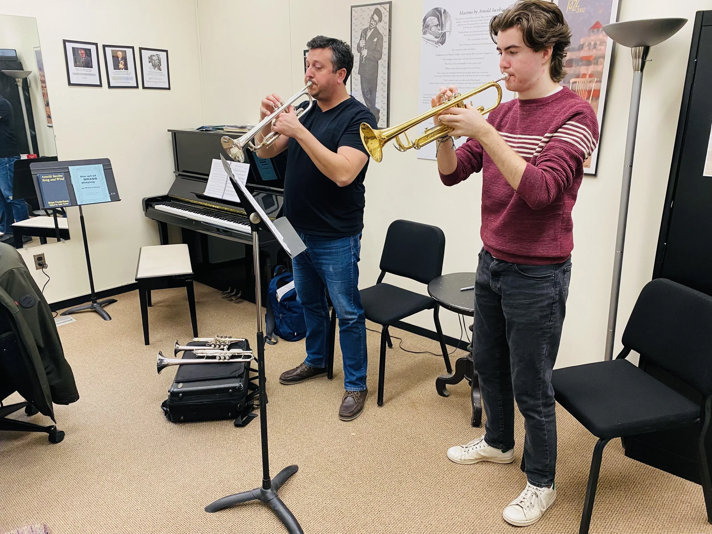 Two men playing trumpets in a music room, with sheet music and a piano nearby, and framed pictures on the wall.