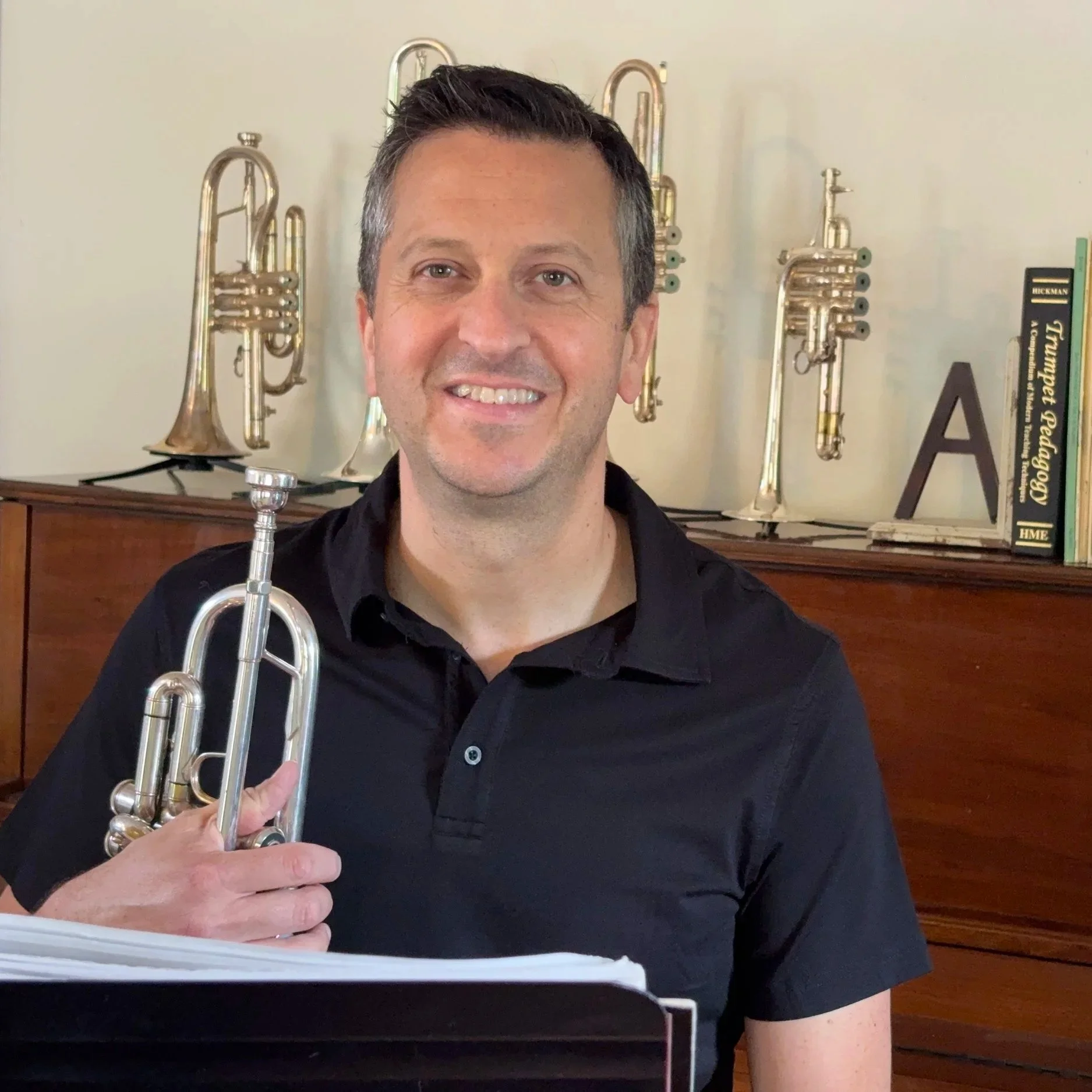 Man smiling at camera holding a brass trumpet, with multiple trumpets on a cabinet and books in the background.