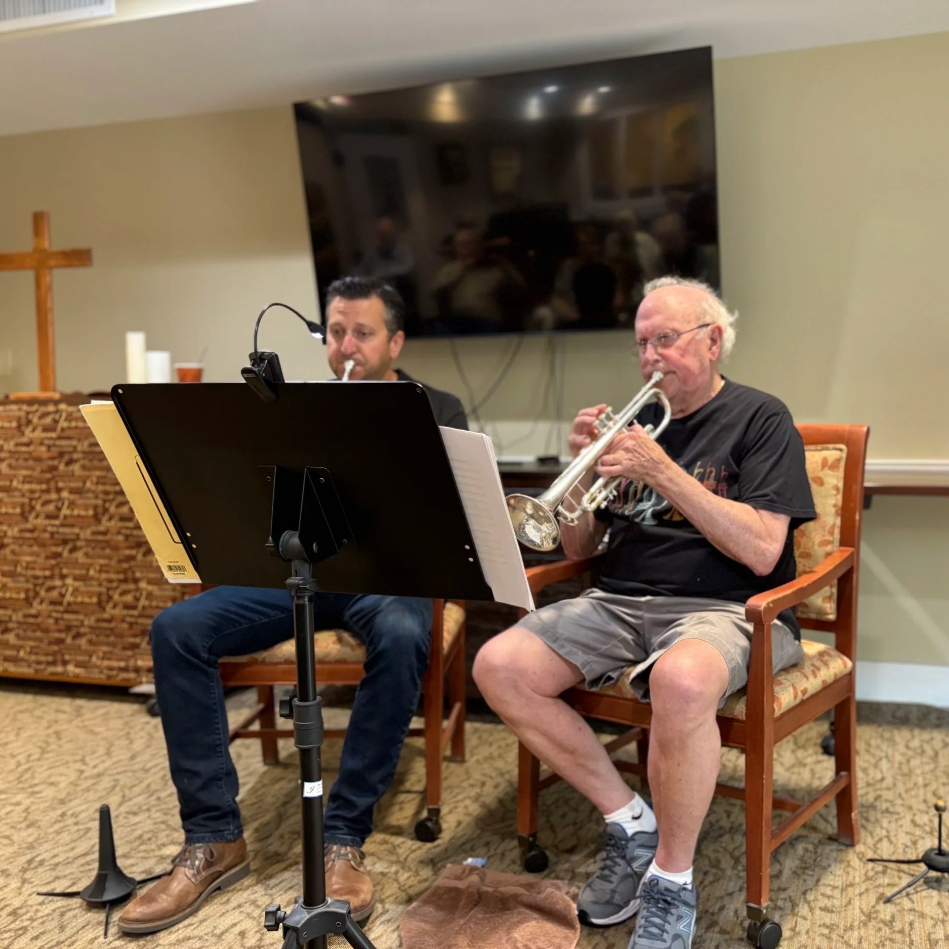 Two men playing musical instruments, one with a trumpet, indoors in a room with a large TV on the wall and a wooden cross decoration in the background.