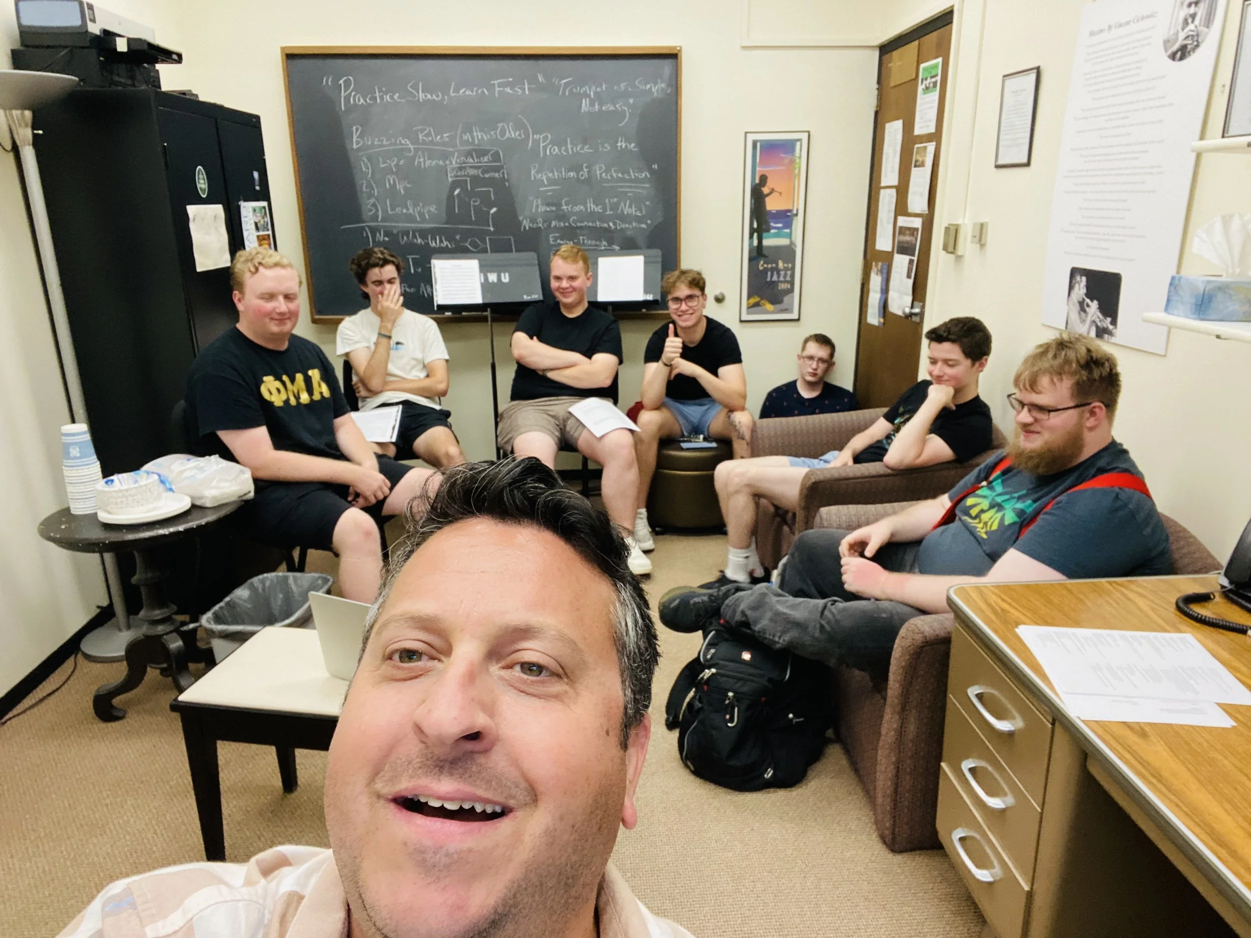 A group of young men sitting and smiling in a classroom or meeting room, with one man taking a selfie. There is a blackboard with handwritten notes in the background, along with posters and papers on the walls.