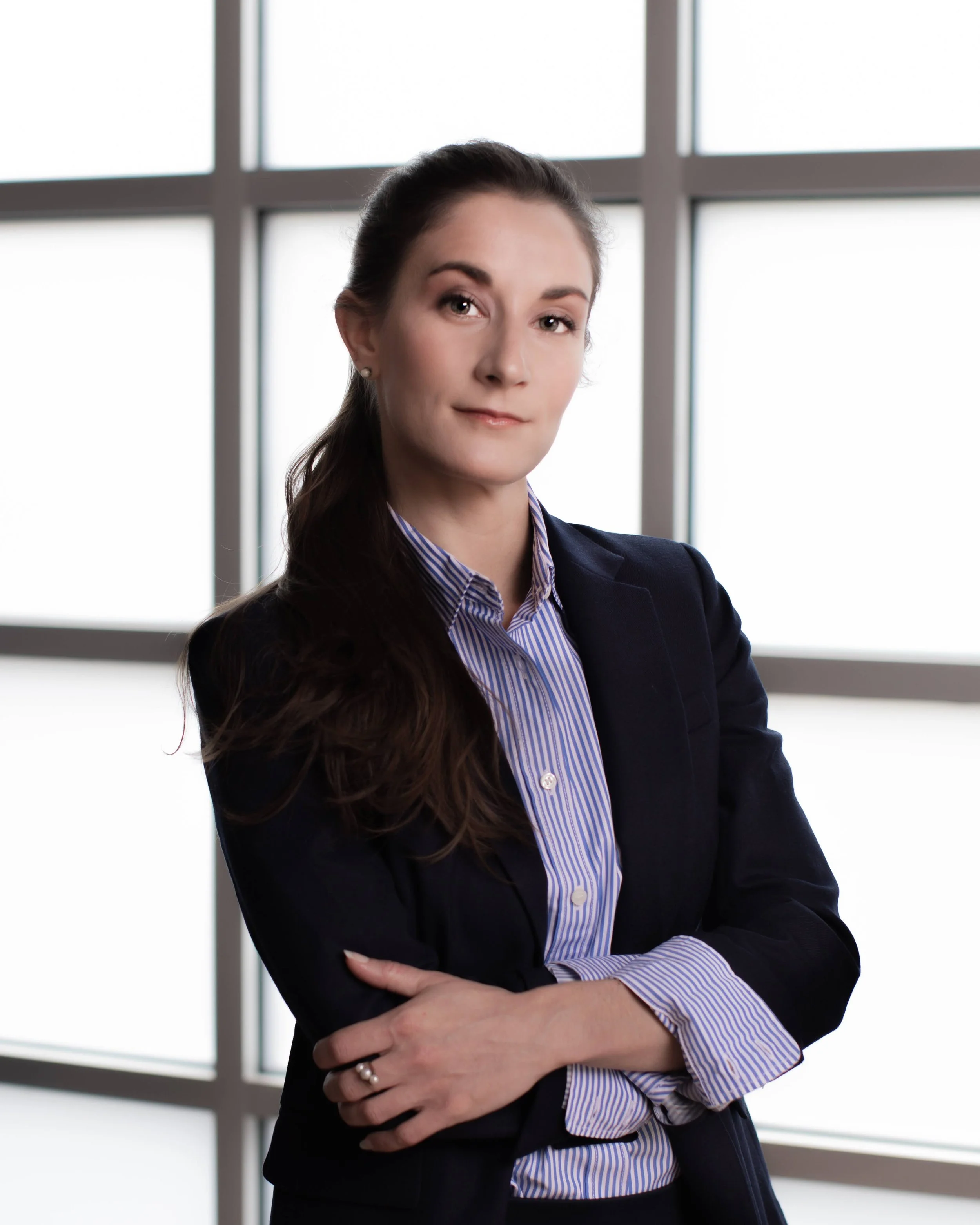 A woman with long brown hair, dressed in a navy blazer and striped shirt, poses with her arms crossed in front of a large window in a professional setting.