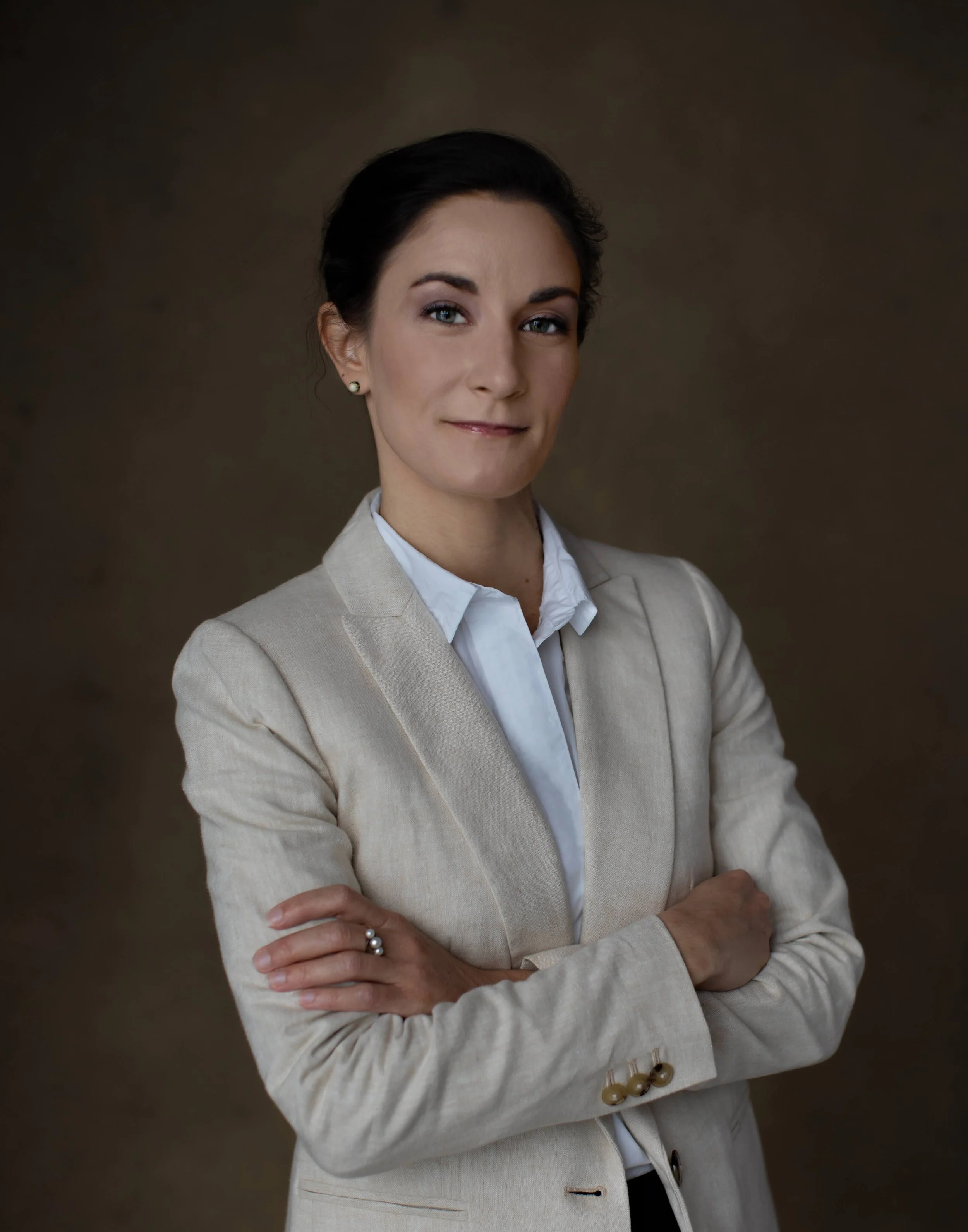 A woman with dark hair in a bun, wearing a beige blazer over a white shirt, standing with arms crossed against a brown background.