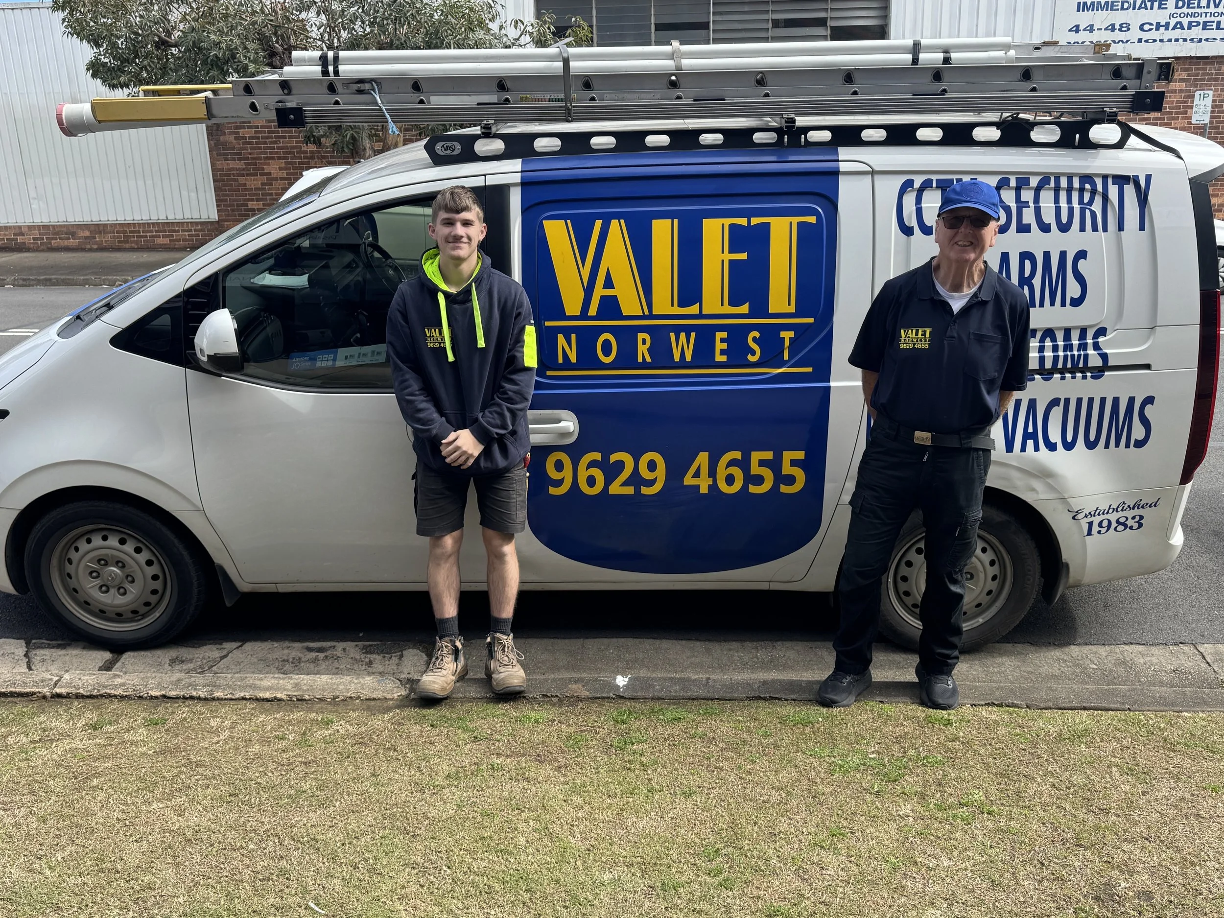 Two men standing next to a white work van with blue and yellow signage that reads "Valet Norwest" and lists services like security, guns, and vacuums. One man is younger, wearing a hoodie and shorts, while the other is older, wearing a uniform and cap. They are posing on a sidewalk with grass in front and buildings in the background.