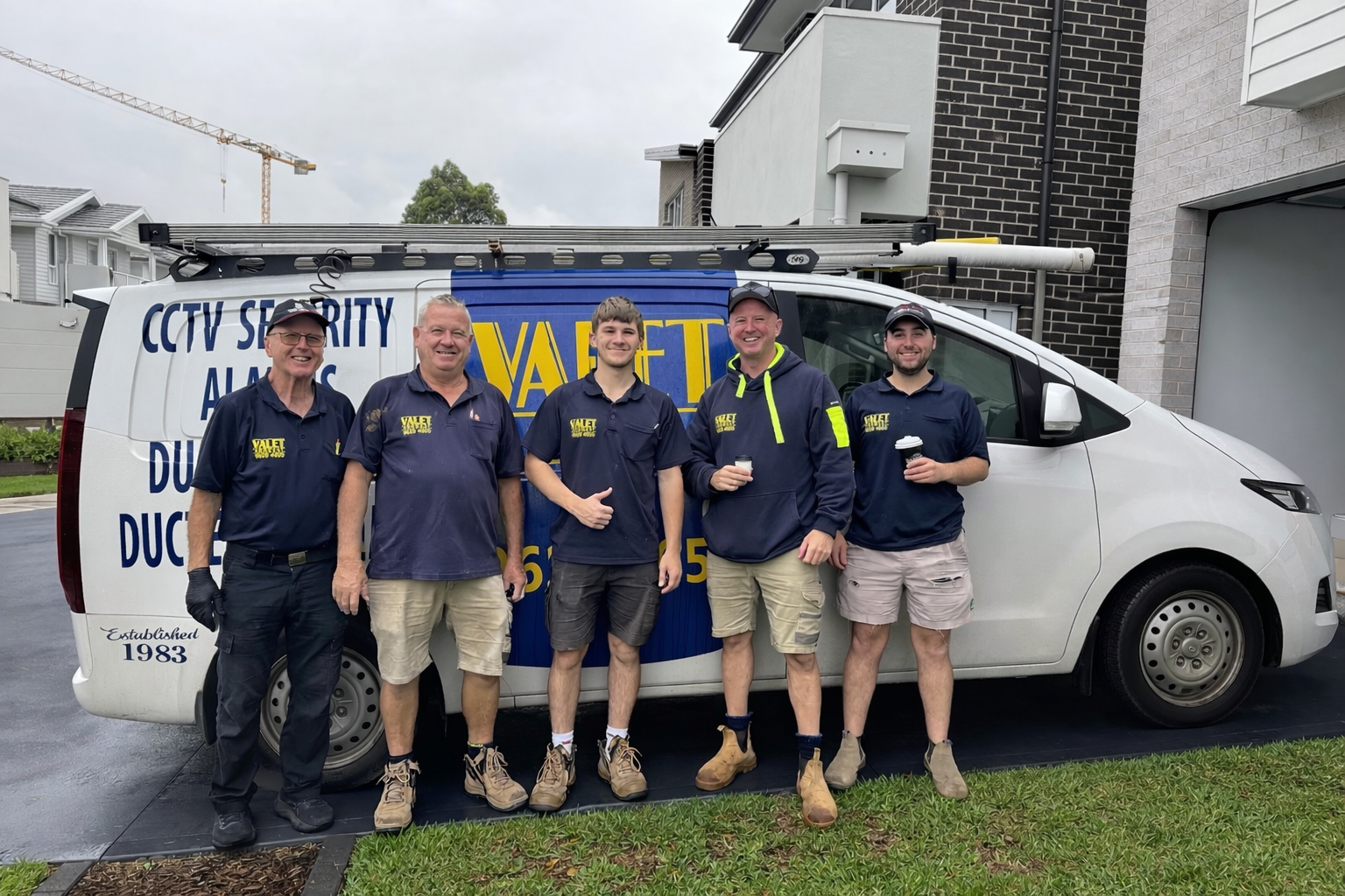 Group of five men in work uniforms standing in front of a company van with ladder on roof, smiling at the camera, holding coffee cups, parked on a driveway with residential buildings in the background.