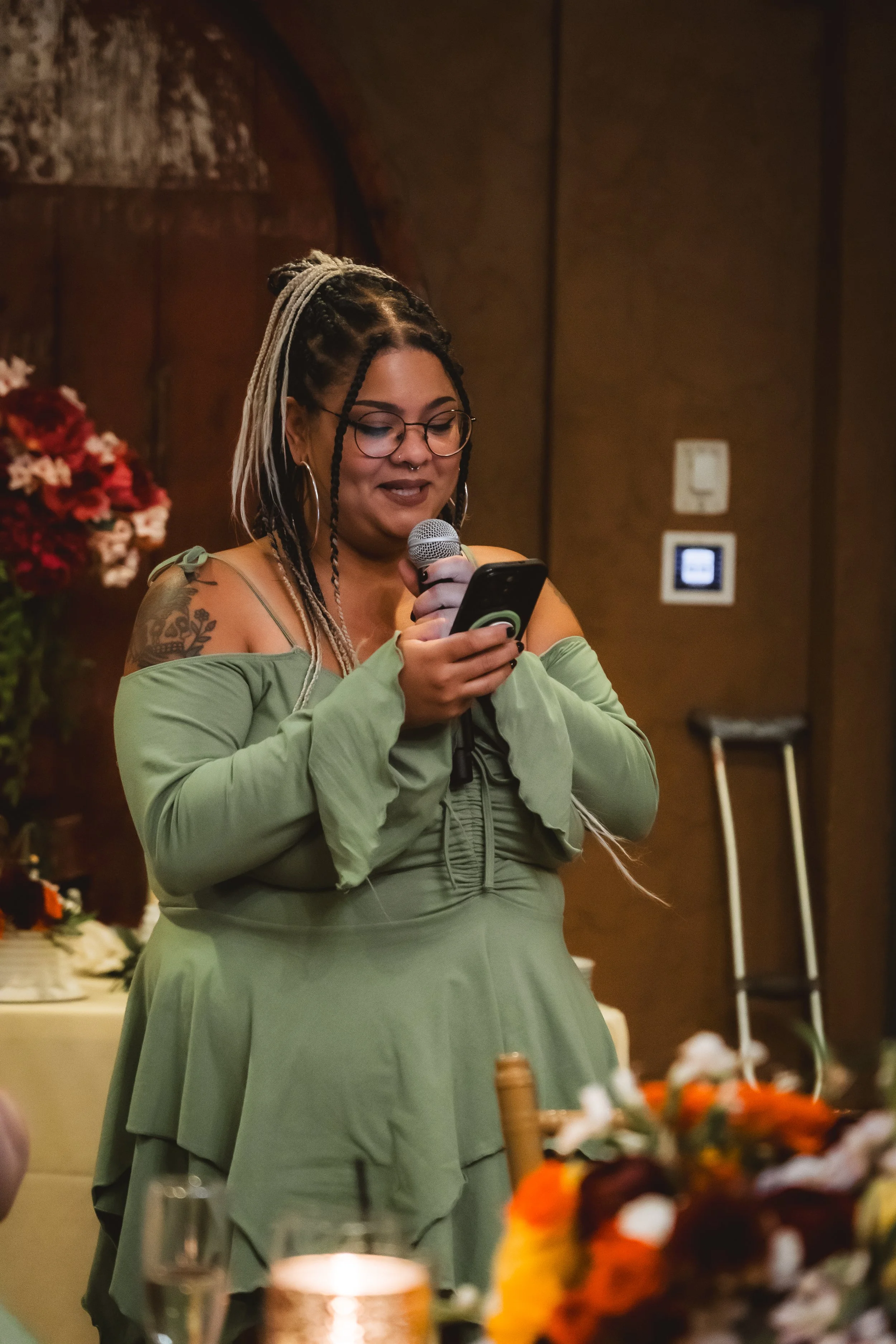 A woman with dreadlocks and glasses standing at a table, reading from her phone into a microphone, during an event in a warmly lit room with floral arrangements.
