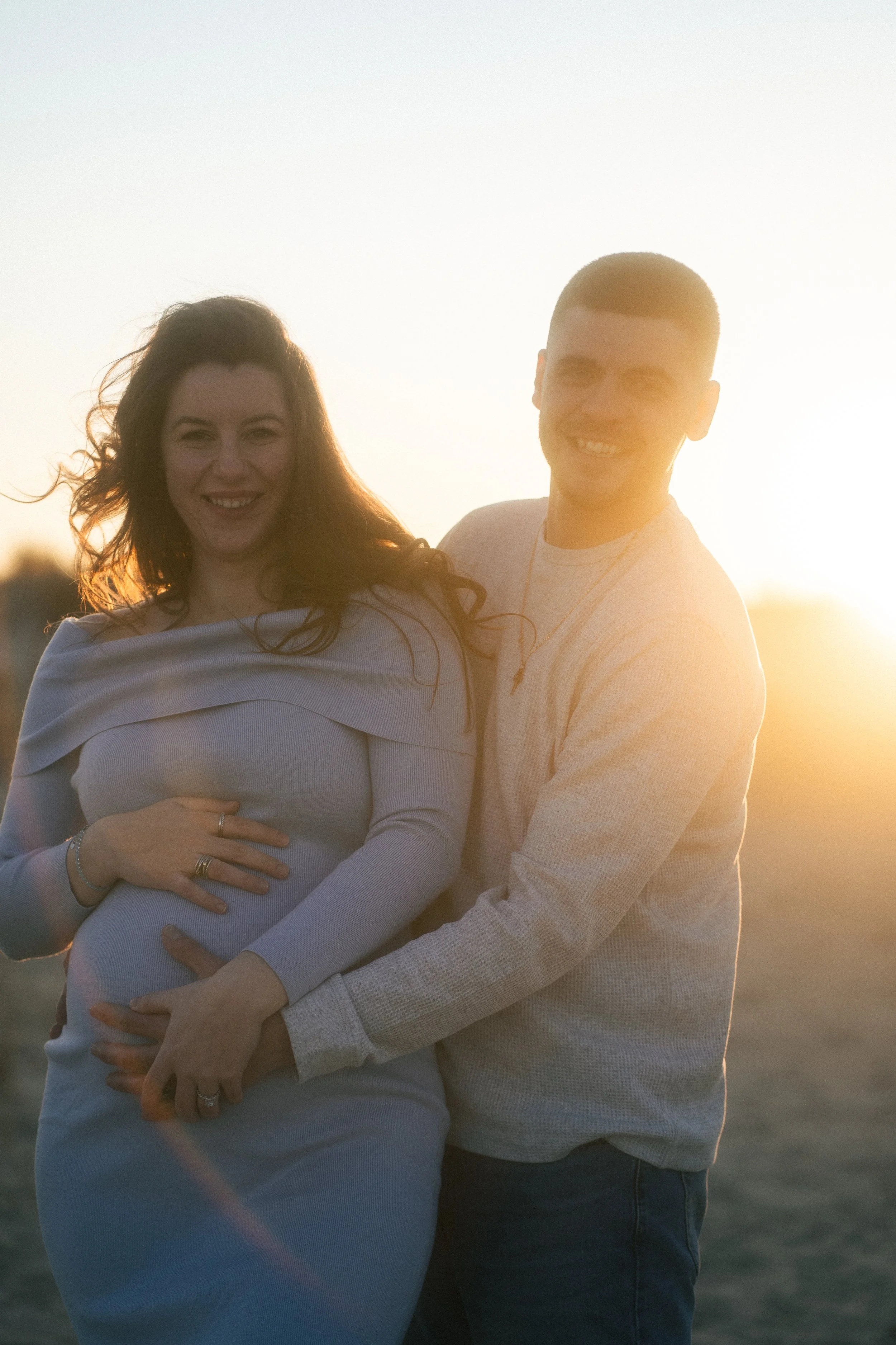 A smiling pregnant woman with dark red hair and a man with short hair standing outdoors during sunset, holding her belly and embracing each other.