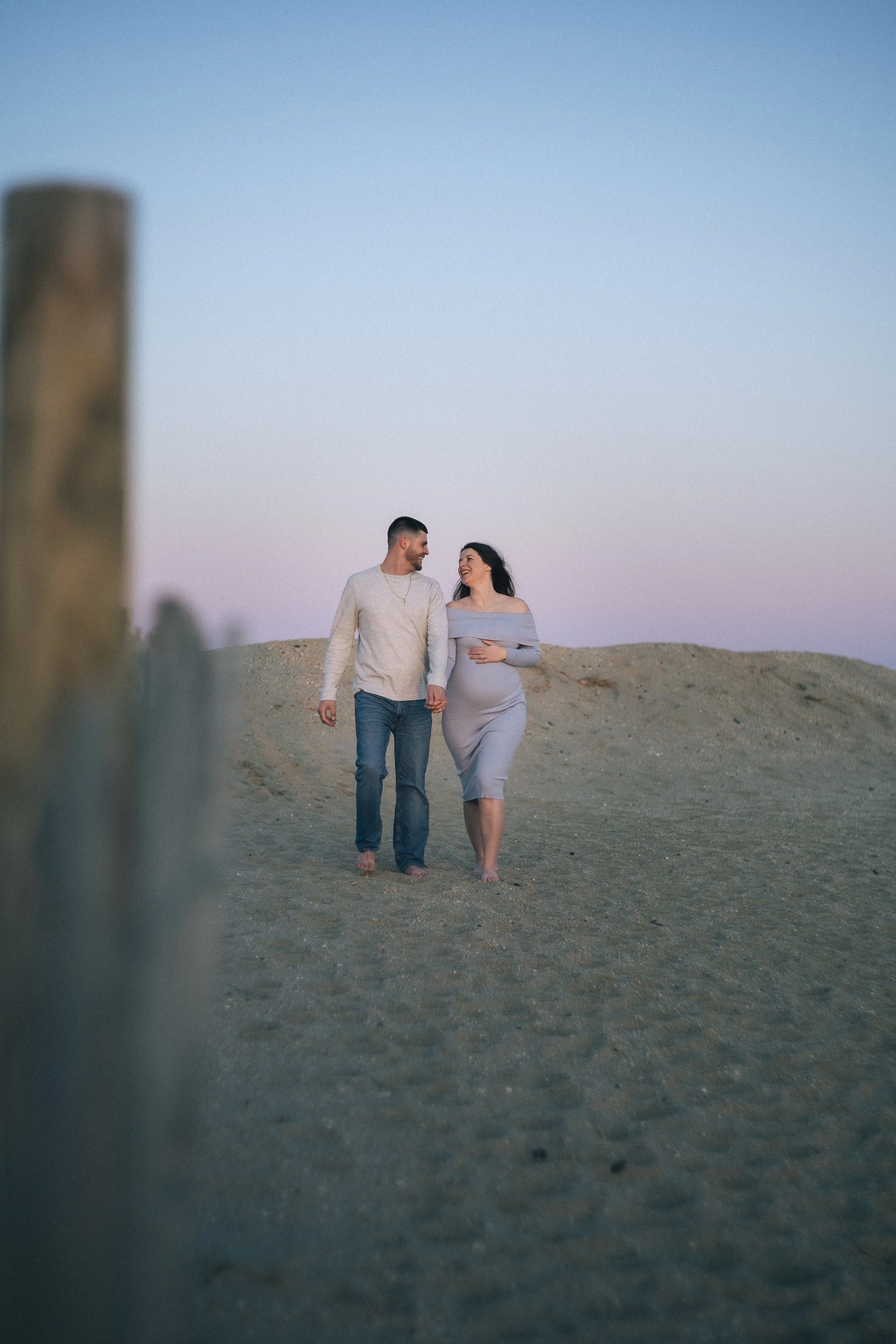 A pregnant woman and man walking on a sandy surface at sunset, smiling and looking at each other.